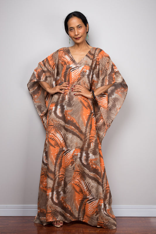 Stylish woman in a floor-length orange and brown abstract kaftan dress with a V-neck and flared sleeves, posed against a gray wall and wooden floor, wearing nude sandals and beaded accessories.

