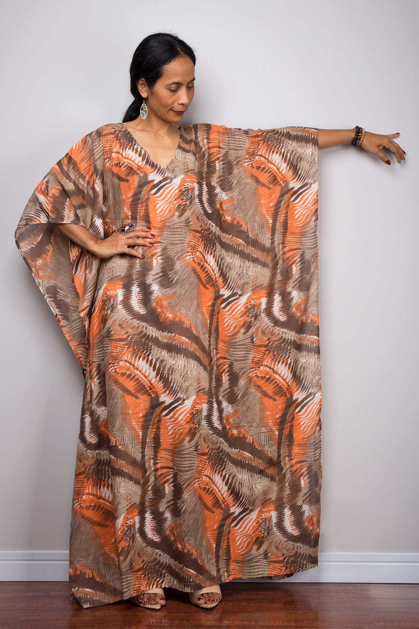 Orange and brown abstract pattern kaftan maxi dress on a woman, featuring a flowing silhouette, V-neck, and short sleeves, set against a minimalist gray and wood background, with turquoise earrings and gold bracelets.

