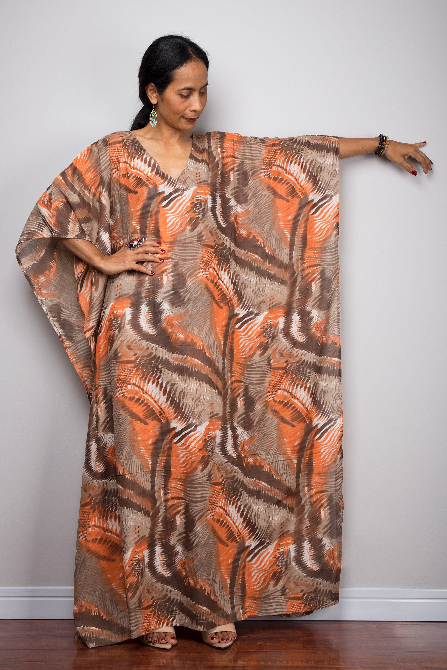 Orange and brown abstract pattern kaftan maxi dress on a woman, featuring a flowing silhouette, V-neck, and short sleeves, set against a minimalist gray and wood background, with turquoise earrings and gold bracelets.

