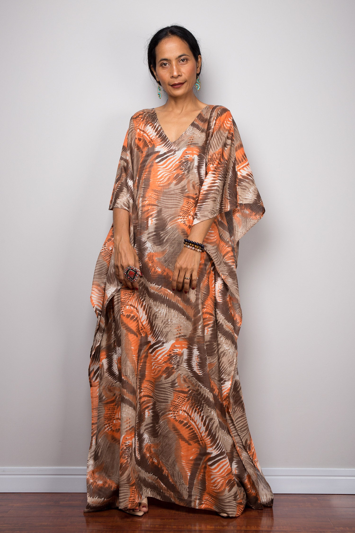 A woman wearing an orange and brown abstract kaftan maxi dress with a V-neckline and wide sleeves, standing against a gray wall with a wooden floor, accessorized with nude sandals, turquoise earrings, beaded bracelets, and a statement ring.

