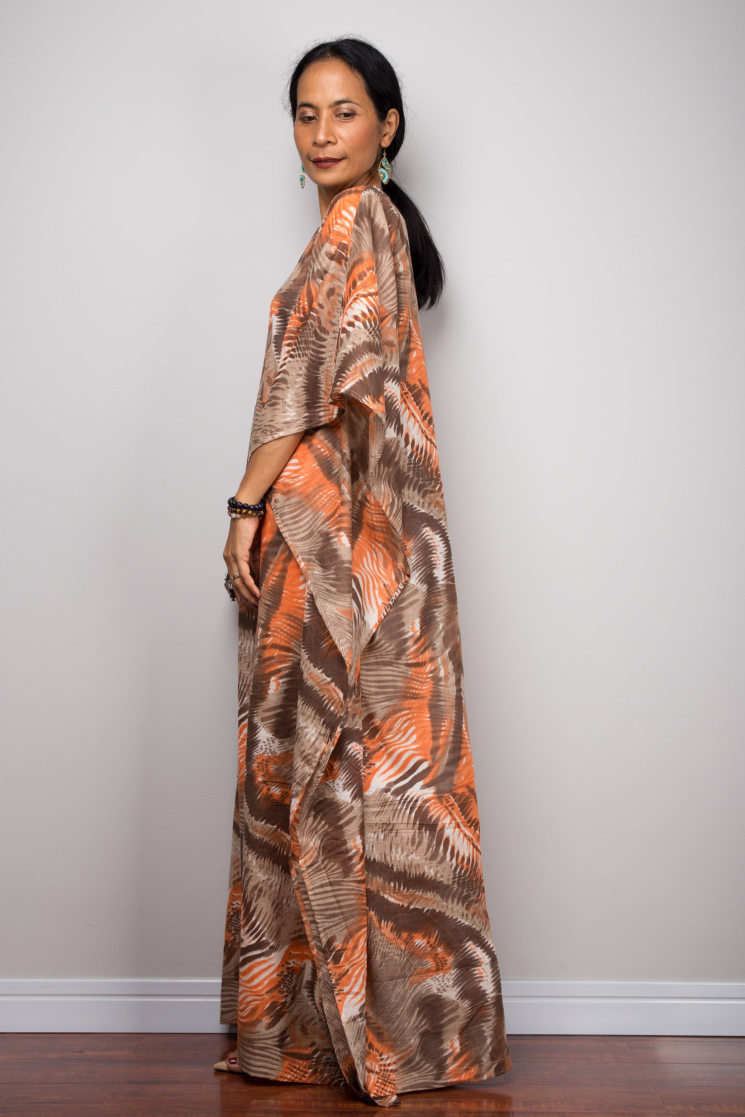 Nuichan orange and brown abstract kaftan maxi dress with a loose fit, deep V-neckline, and wide sleeves, modeled by a woman with turquoise earrings and bracelets, against a simple gray and wood backdrop.

