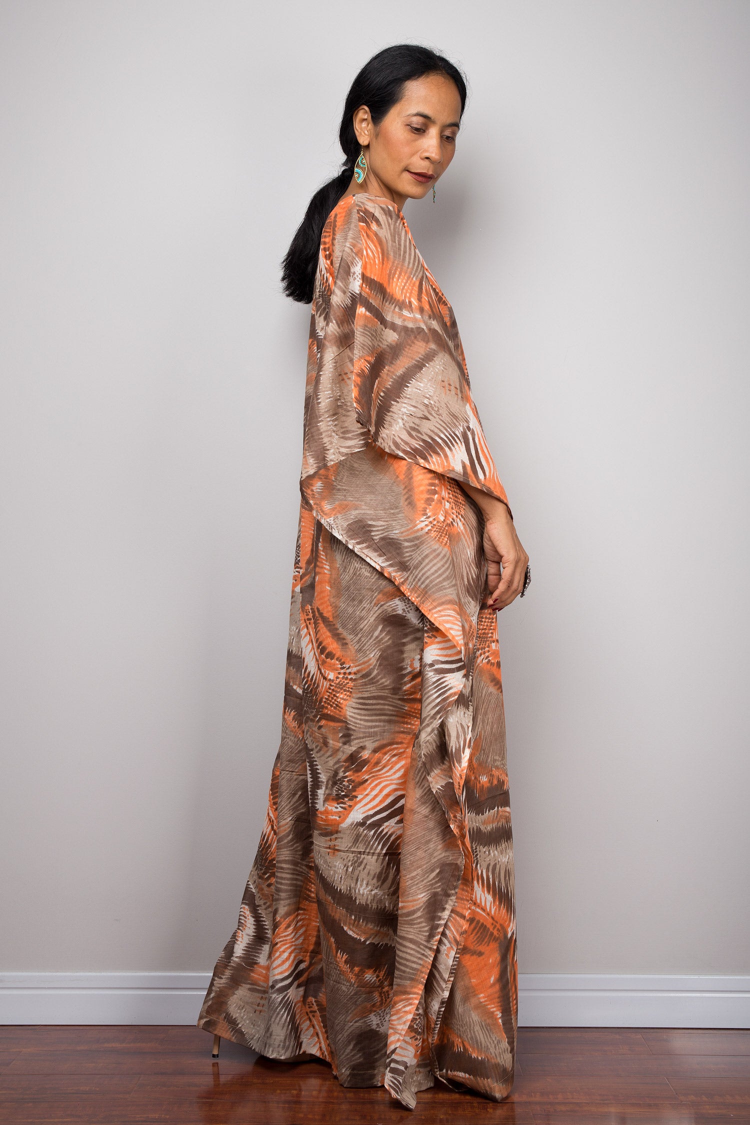 Elegant orange and brown abstract kaftan maxi dress with a V-neck and wide sleeves, worn by a woman against a gray wall and wooden floor, paired with nude sandals, turquoise earrings, and statement jewelry.

