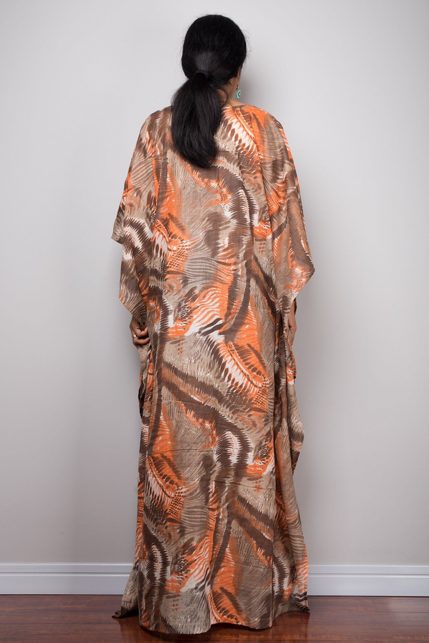 Model in an orange and brown abstract pattern kaftan maxi dress featuring a deep V-neck and flared short sleeves, standing on a wooden floor with a gray wall background, accessorized with nude sandals and beaded bracelets.

