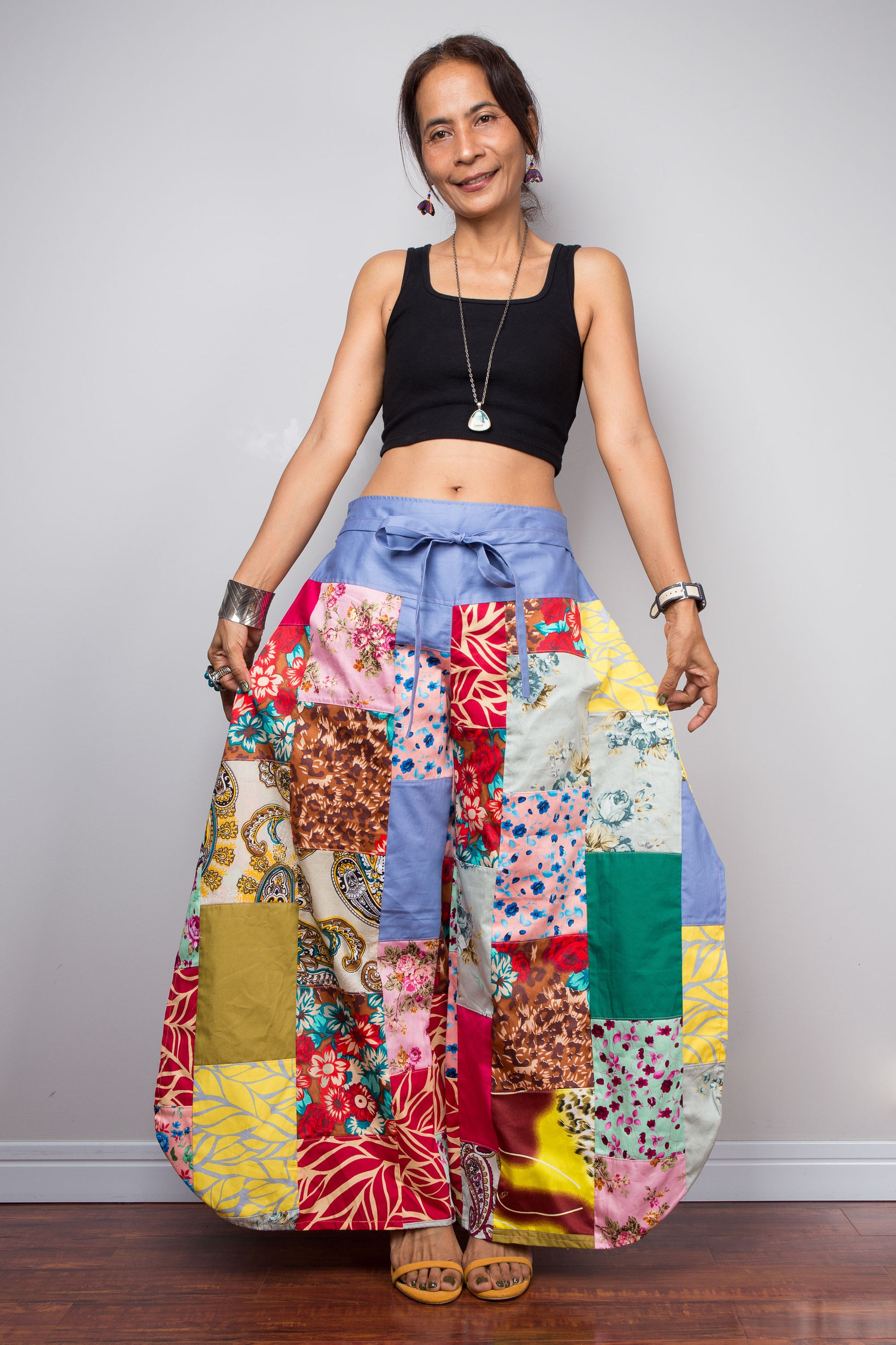 Front-facing view of a petite model holding out the wide legs of the Nuichan patchwork palazzo pants to display the full maximalist quilt-style patchwork construction, periwinkle blue solid waistband with self-tie sash bow at the front, and mixed cotton satin print panels in red, pink, yellow, teal, green, paisley and floral patterns