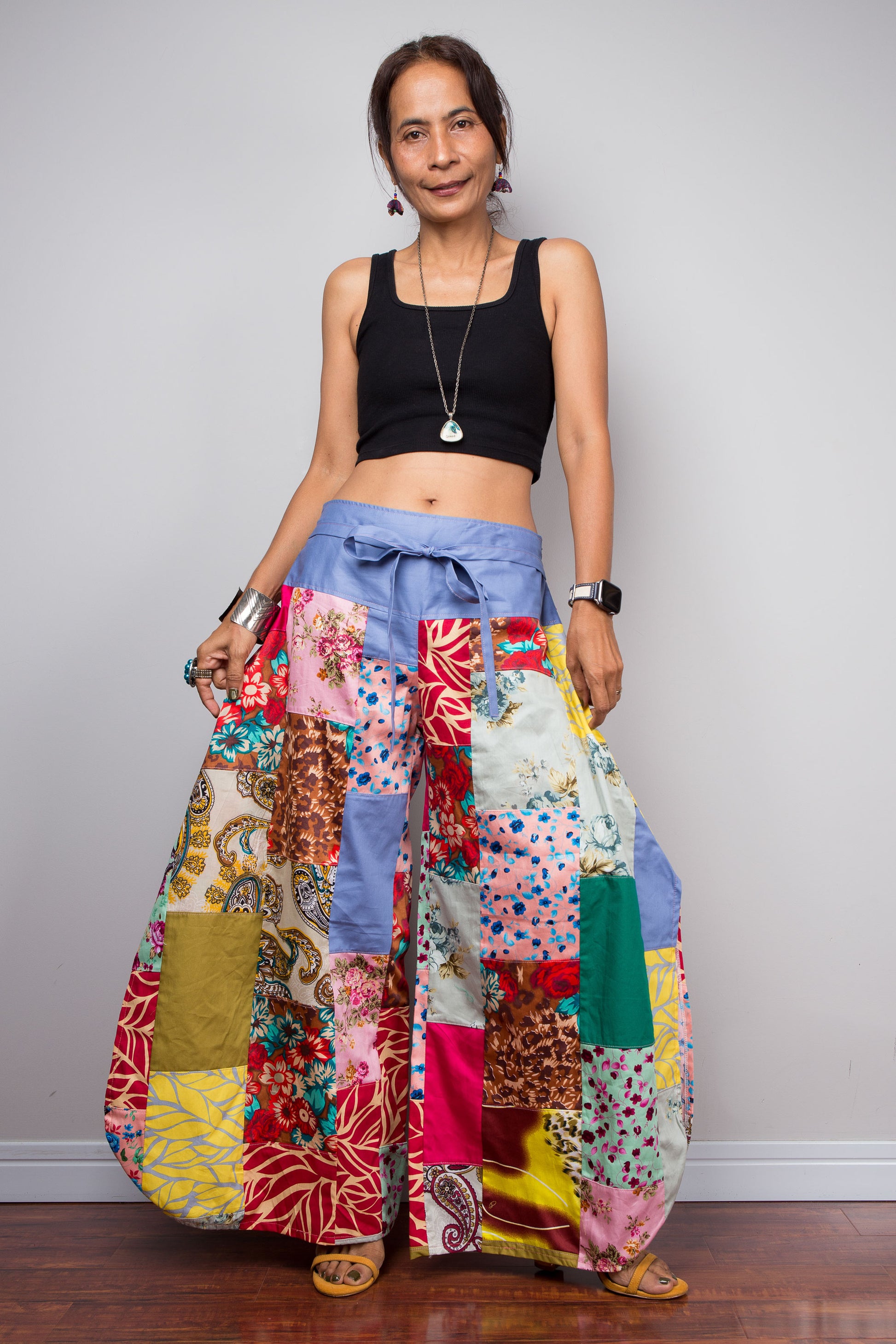 Front-facing view of a petite female model holding out the wide legs of the Nuichan patchwork palazzo pants to display the full maximalist quilt-style patchwork construction, periwinkle blue solid waistband with self-tie sash bow at the front, and mixed cotton satin print panels in red, pink, yellow, teal, green, paisley and floral patterns