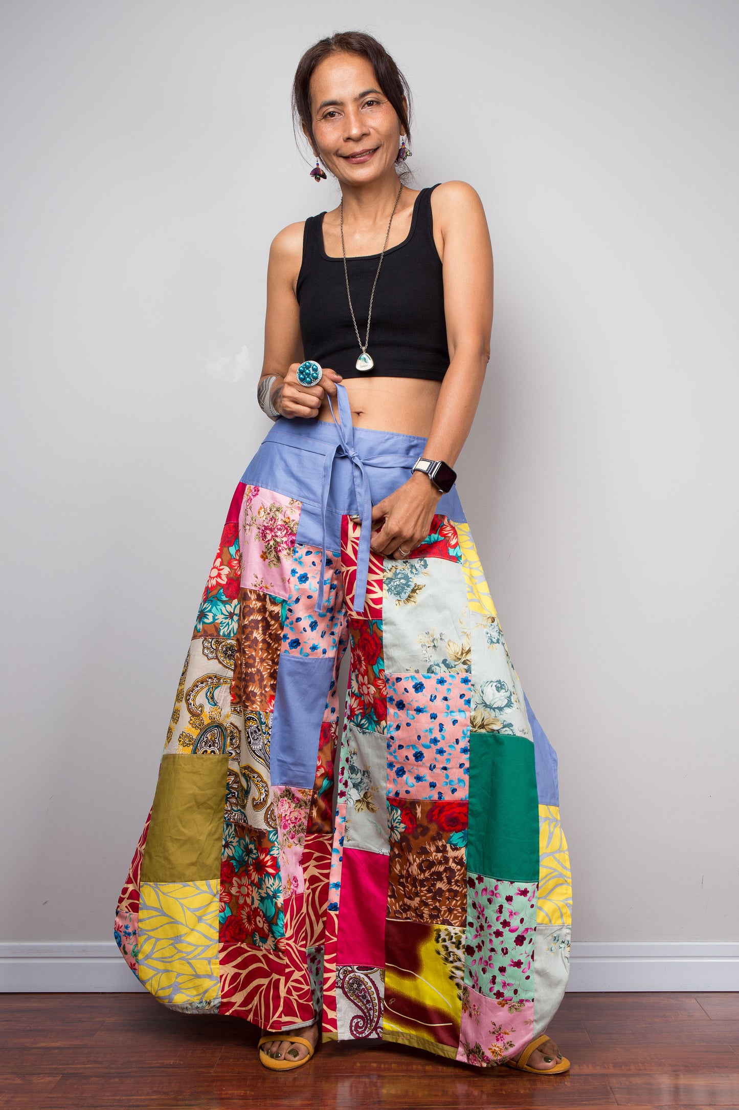 Front-facing view of a model holding out the wide legs of the Nuichan patchwork palazzo pants to display the full maximalist quilt-style patchwork construction, periwinkle blue solid waistband with self-tie sash bow at the front, and mixed cotton print panels in red, pink, yellow, teal, green, paisley and floral patterns
