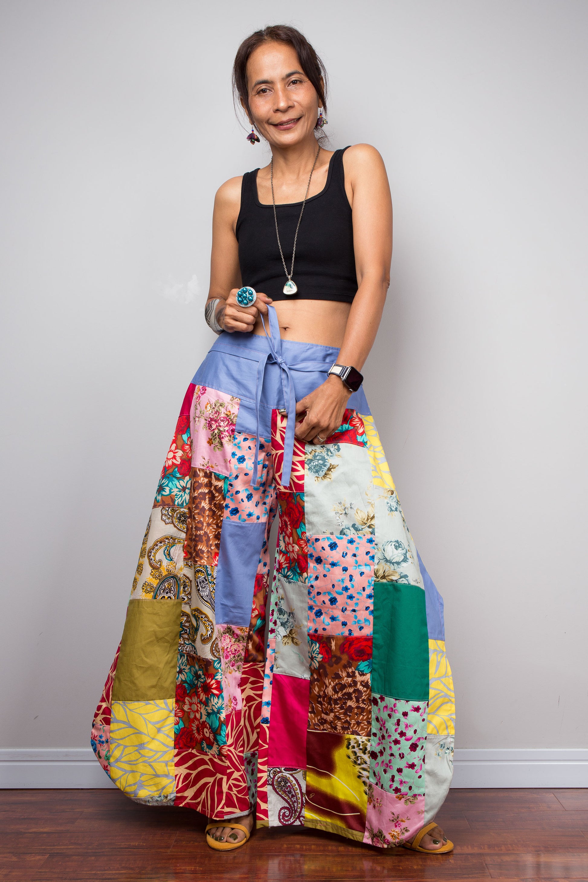Front-facing view of a model holding out the wide legs of the Nuichan patchwork palazzo pants to display the full maximalist quilt-style patchwork construction, periwinkle blue solid waistband with self-tie sash bow at the front, and mixed cotton print panels in red, pink, yellow, teal, green, paisley and floral patterns