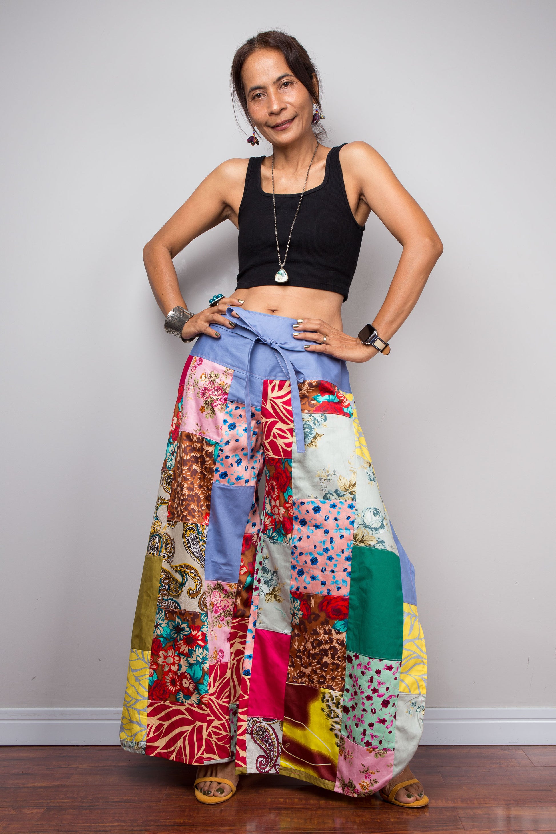 Front-facing view of a model holding hand on hips whilst wearing the Nuichan patchwork palazzo pants to display the full maximalist quilt-style patchwork construction, periwinkle blue solid waistband with self-tie sash bow at the front, and mixed cotton satin print panels in red, pink, yellow, teal, green, paisley and floral patterns