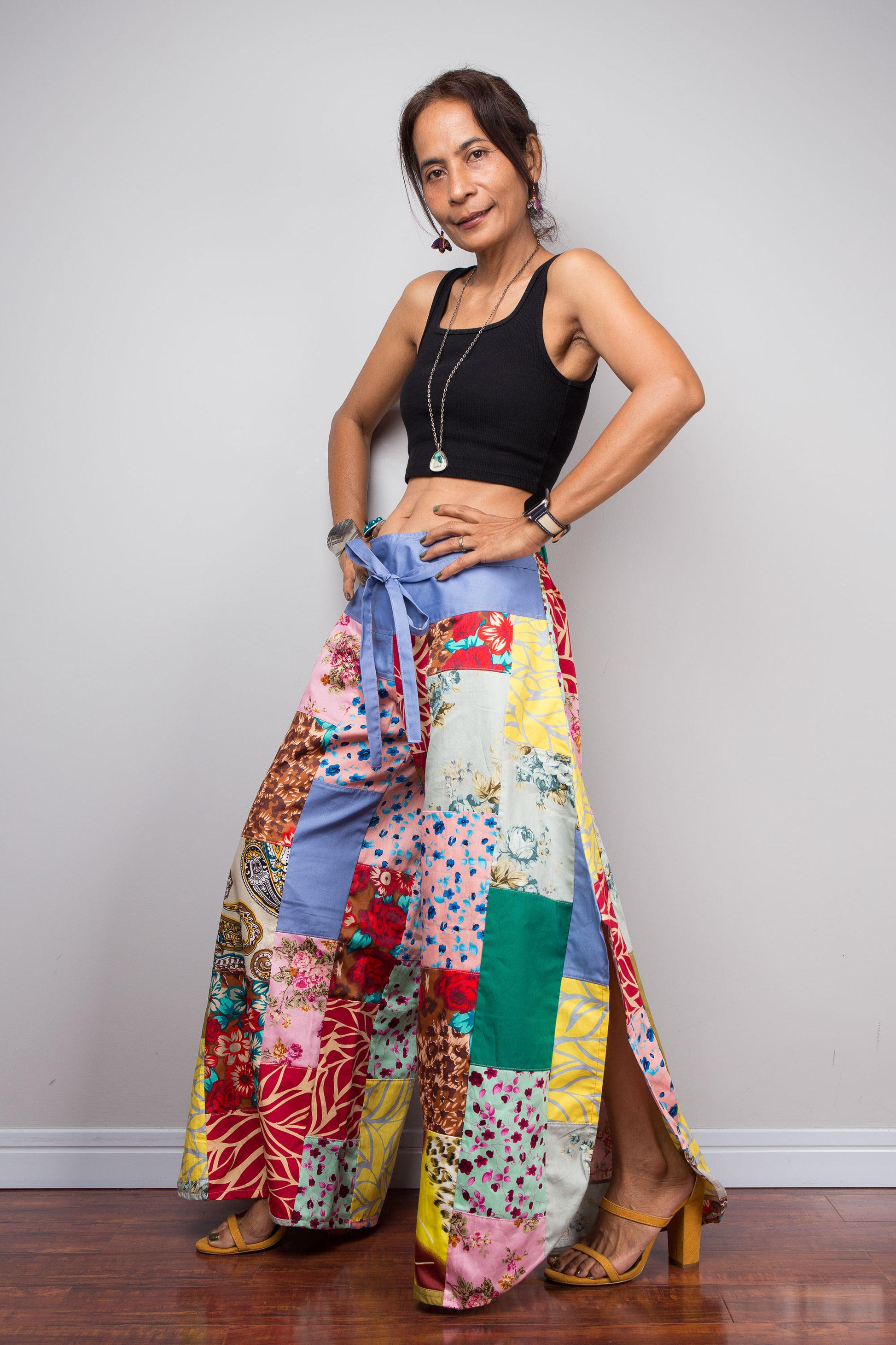 Three quarter front view of a model holding out the wide legs of the Nuichan patchwork palazzo pants to display the full maximalist quilt-style patchwork construction, periwinkle blue solid waistband with self-tie sash bow at the front, and mixed cotton satin print panels in red, pink, yellow, teal, green, paisley and floral patterns