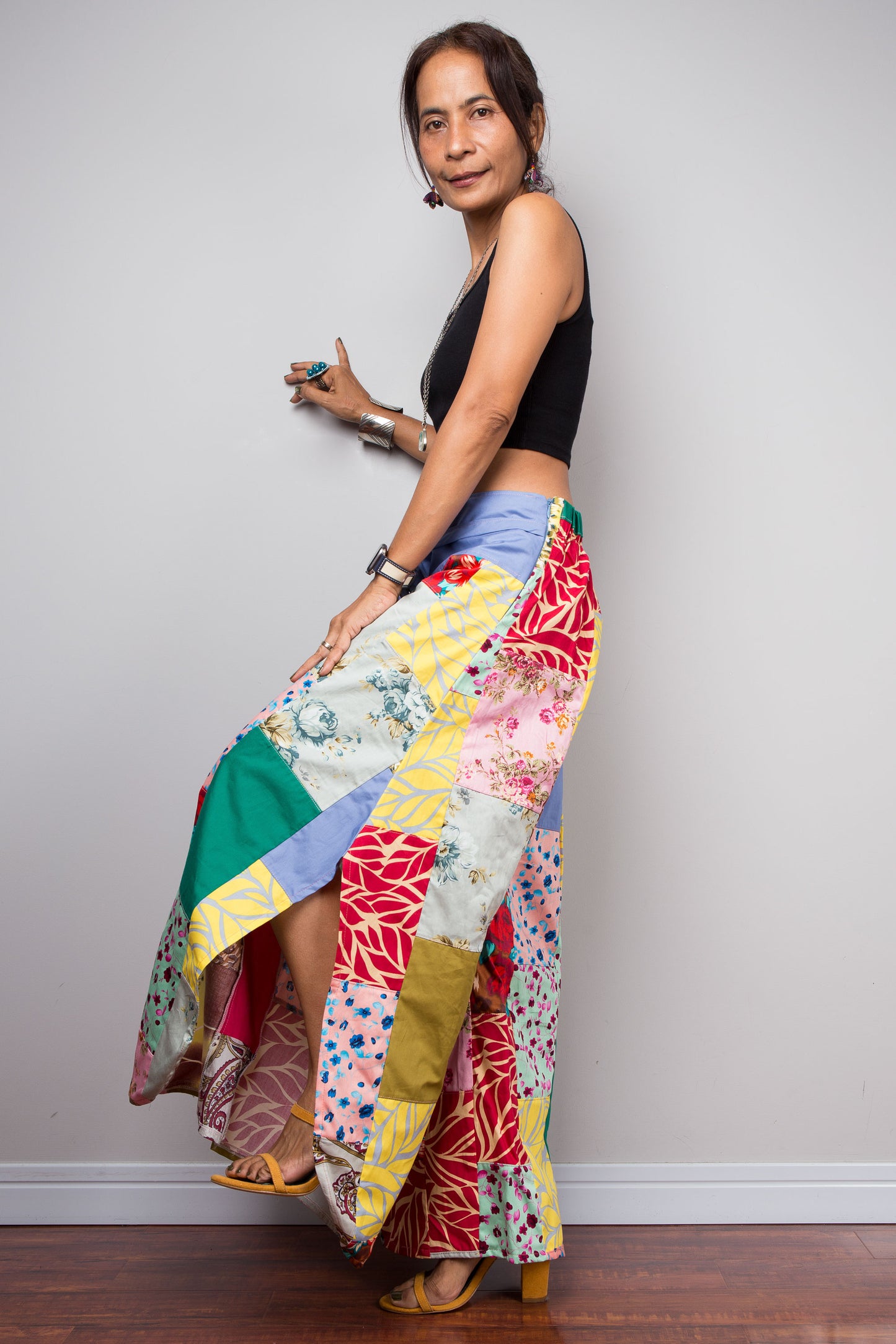 Side view of a model holding out the wide legs of the Nuichan patchwork palazzo pants to display the full maximalist quilt-style patchwork construction, periwinkle blue solid waistband with self-tie sash bow at the front, and mixed cotton satin print panels in red, pink, yellow, teal, green, paisley and floral patterns