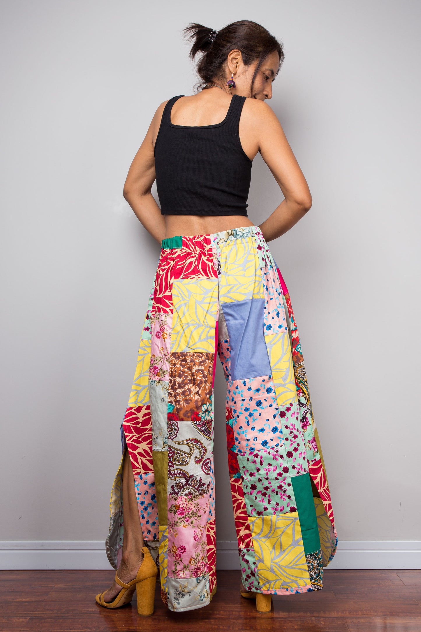 Rear view of a model wearing the Nuichan handmade patchwork wide leg palazzo pants, displaying the full ultra-wide leg volume and hand-sewn quilt-style patchwork construction in mixed cotton satin prints including florals, botanicals, paisley, teal and yellow leaf prints, with side split detail visible at the hem