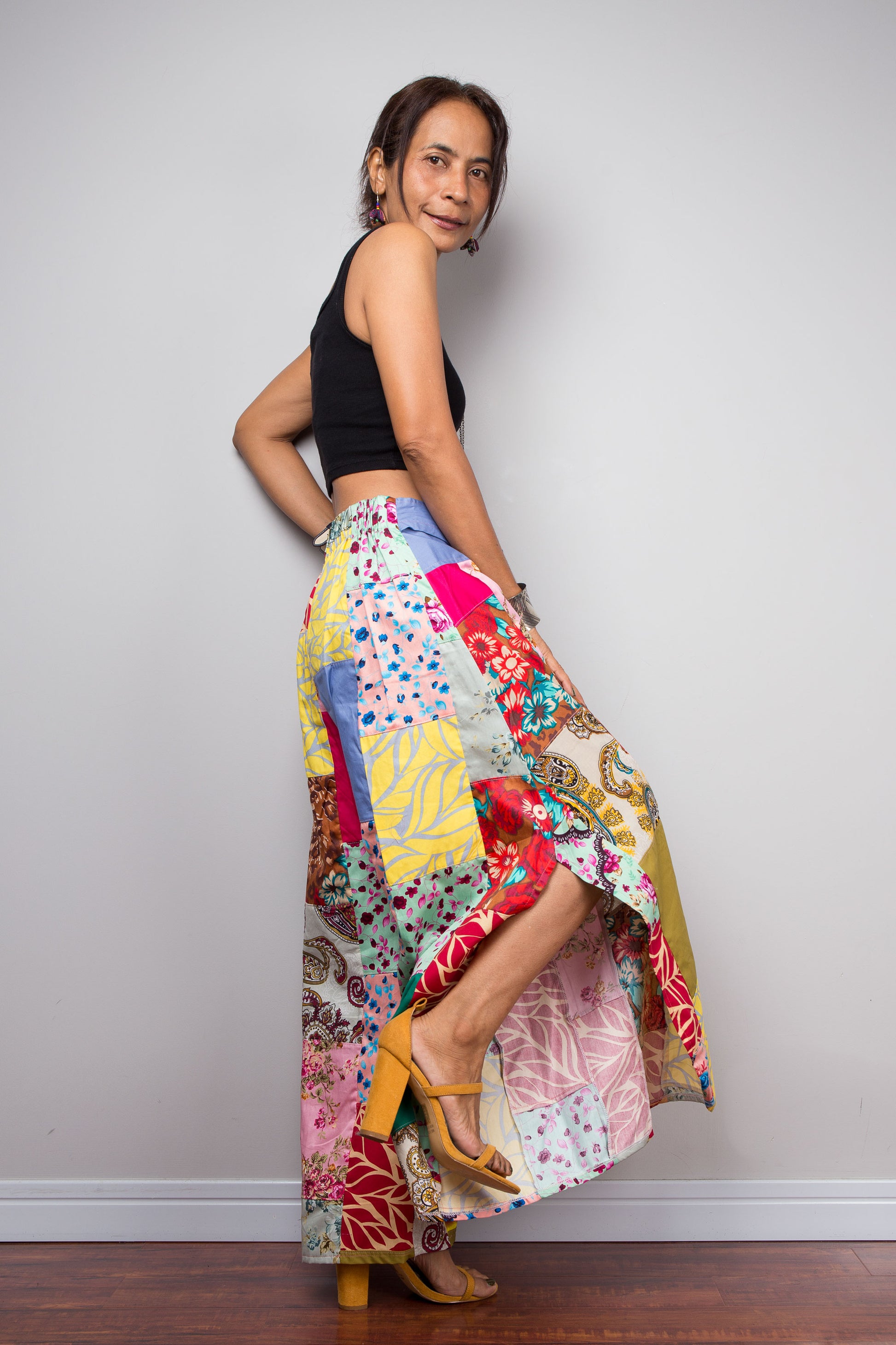 Side facing view of a model holding out the wide legs of the Nuichan patchwork palazzo pants to display the full maximalist quilt-style patchwork construction, periwinkle blue solid waistband with self-tie sash bow at the front, and mixed cotton satin print panels in red, pink, yellow, teal, green, paisley and floral patterns