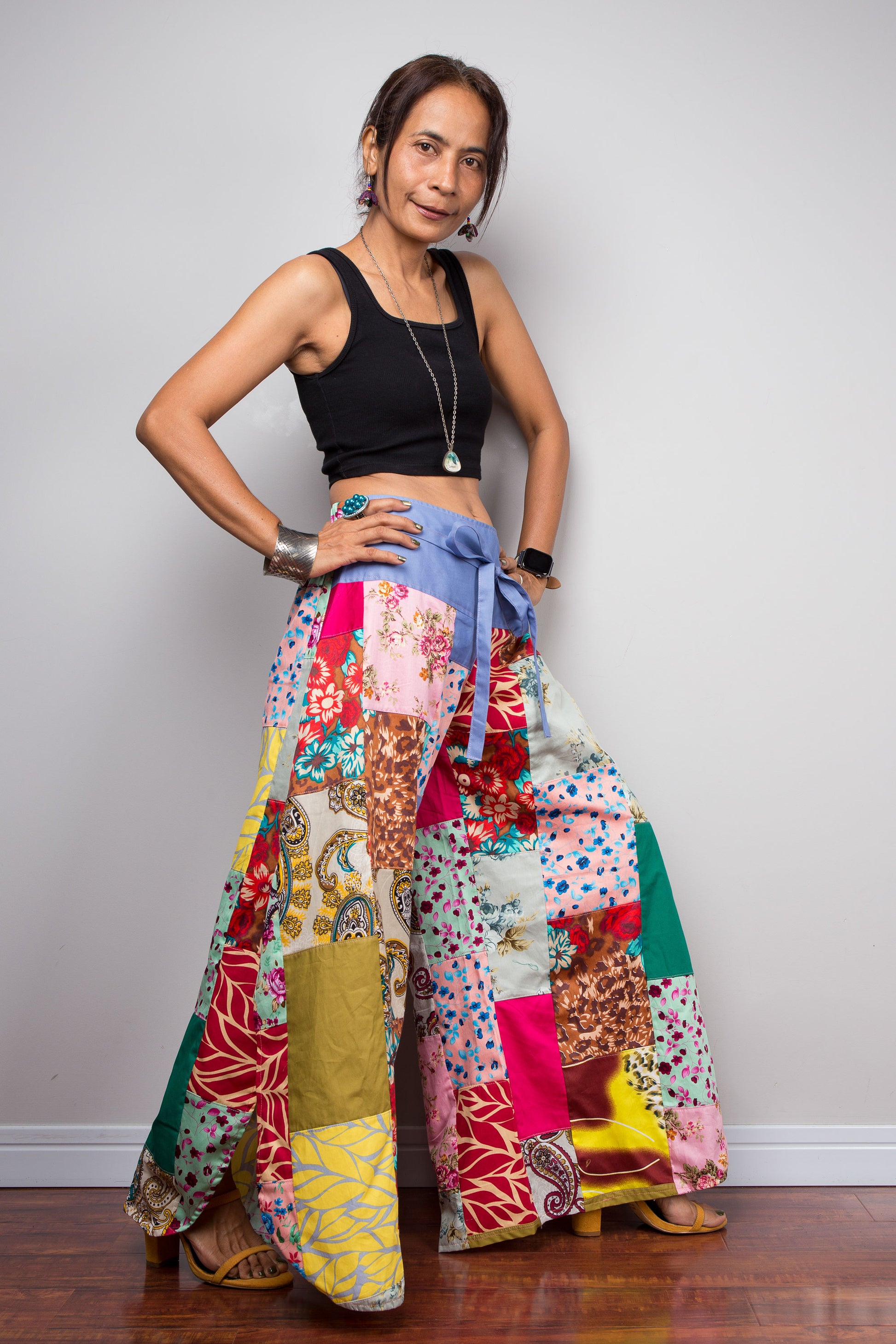 Three-quarter side view of a model wearing the Nuichan handmade cotton satin patchwork wide leg palazzo pants with hands on hips, showcasing the periwinkle blue self-tie sash waistband, dramatic ultra-wide leg silhouette, side split detail at the hem, and the full maximalist mixed print patchwork panel construction