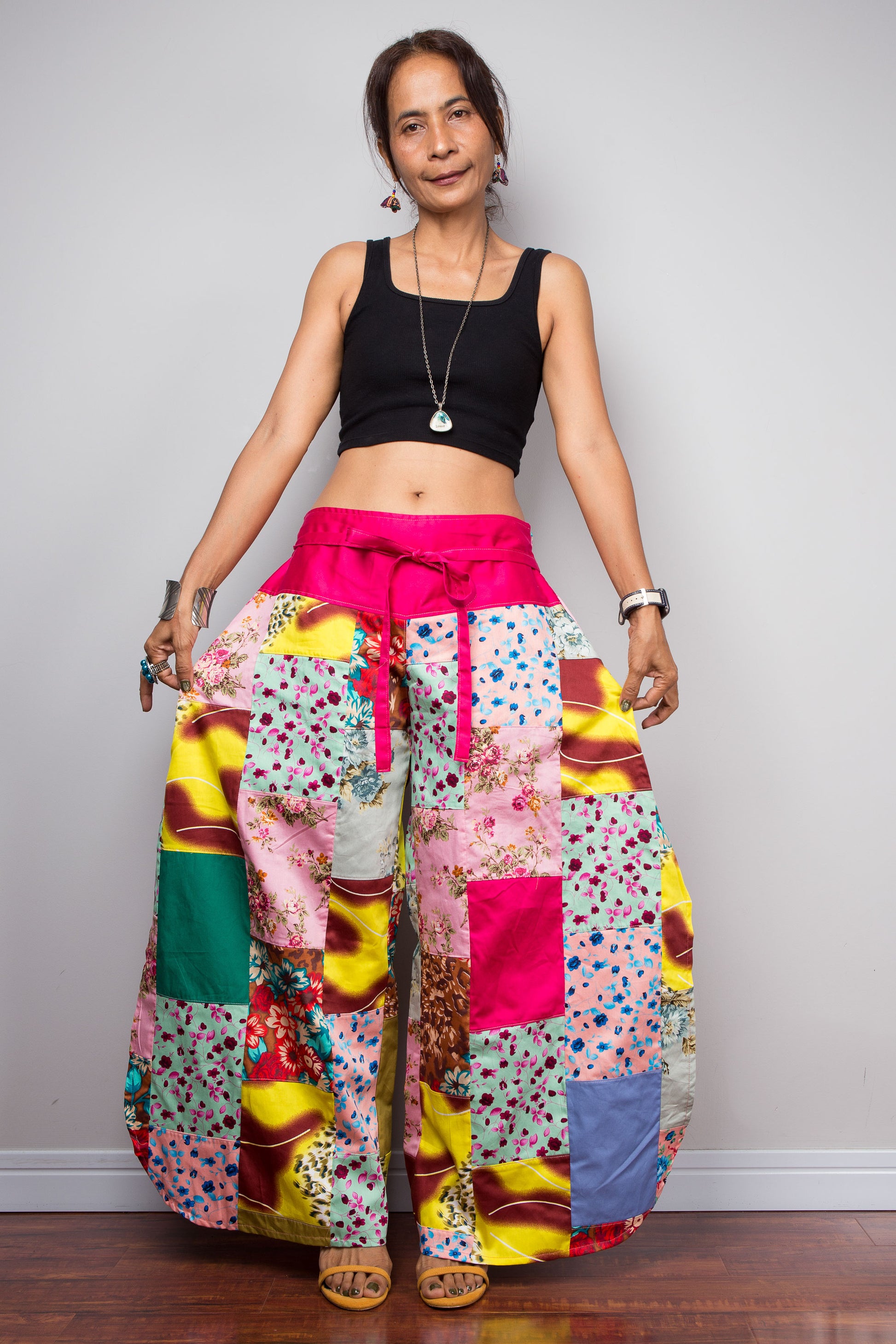 Front-facing view of a model wearing the Nuichan pastel patchwork wide leg palazzo pants, showcasing the vivid pink self-tie sash waistband, full ultra-wide leg silhouette, and hand-sewn quilt-style patchwork construction in mixed cotton satin pastel prints including mint, blush pink, teal, yellow satin and floral panels, styled with a black crop top