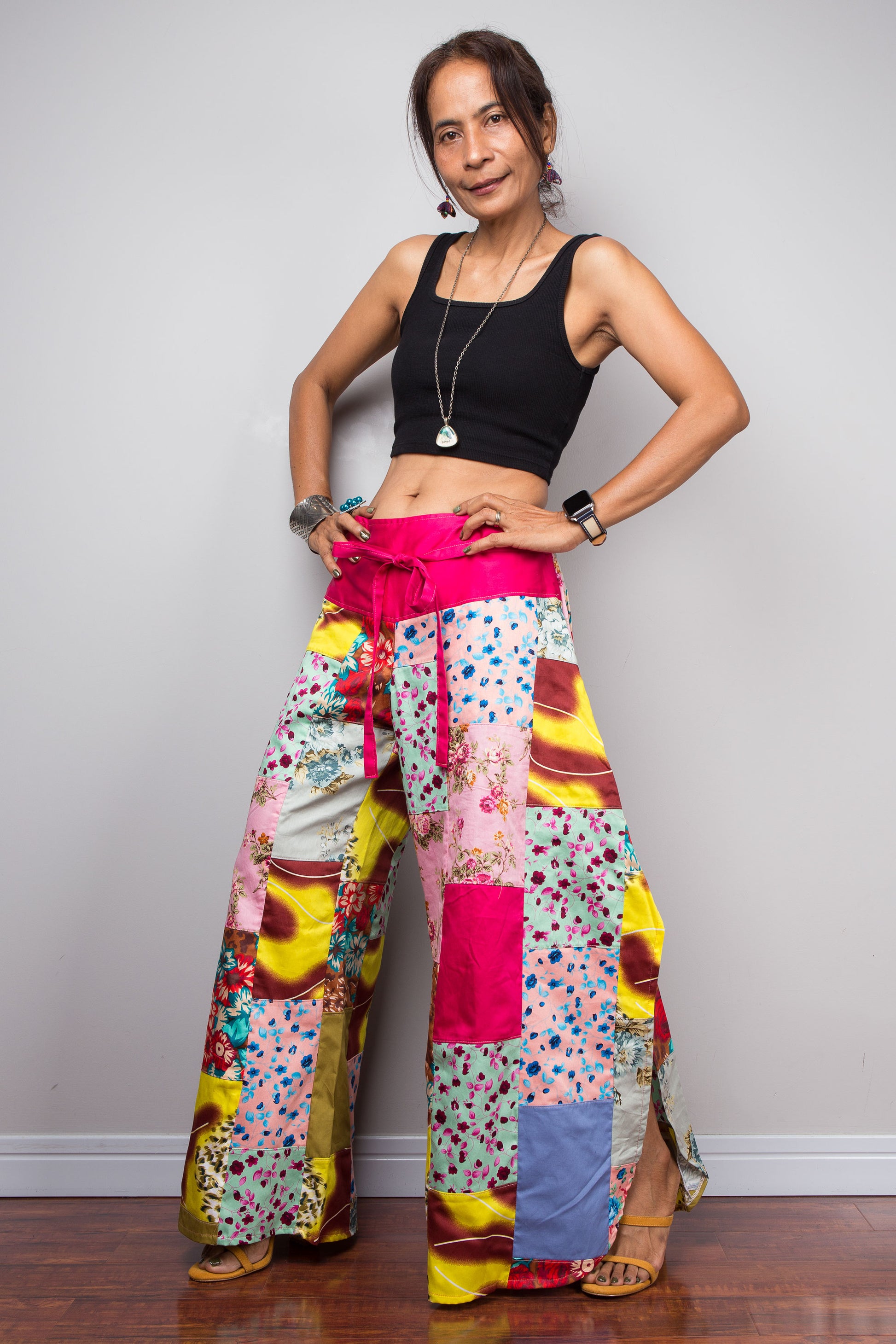 Three quarter view of a model wearing the Nuichan pastel patchwork wide leg palazzo pants, showcasing the vivid pink self-tie sash waistband, full ultra-wide leg silhouette, and hand-sewn quilt-style patchwork construction in mixed cotton satin pastel prints including mint, blush pink, teal, yellow satin and floral panels, styled with a black crop top