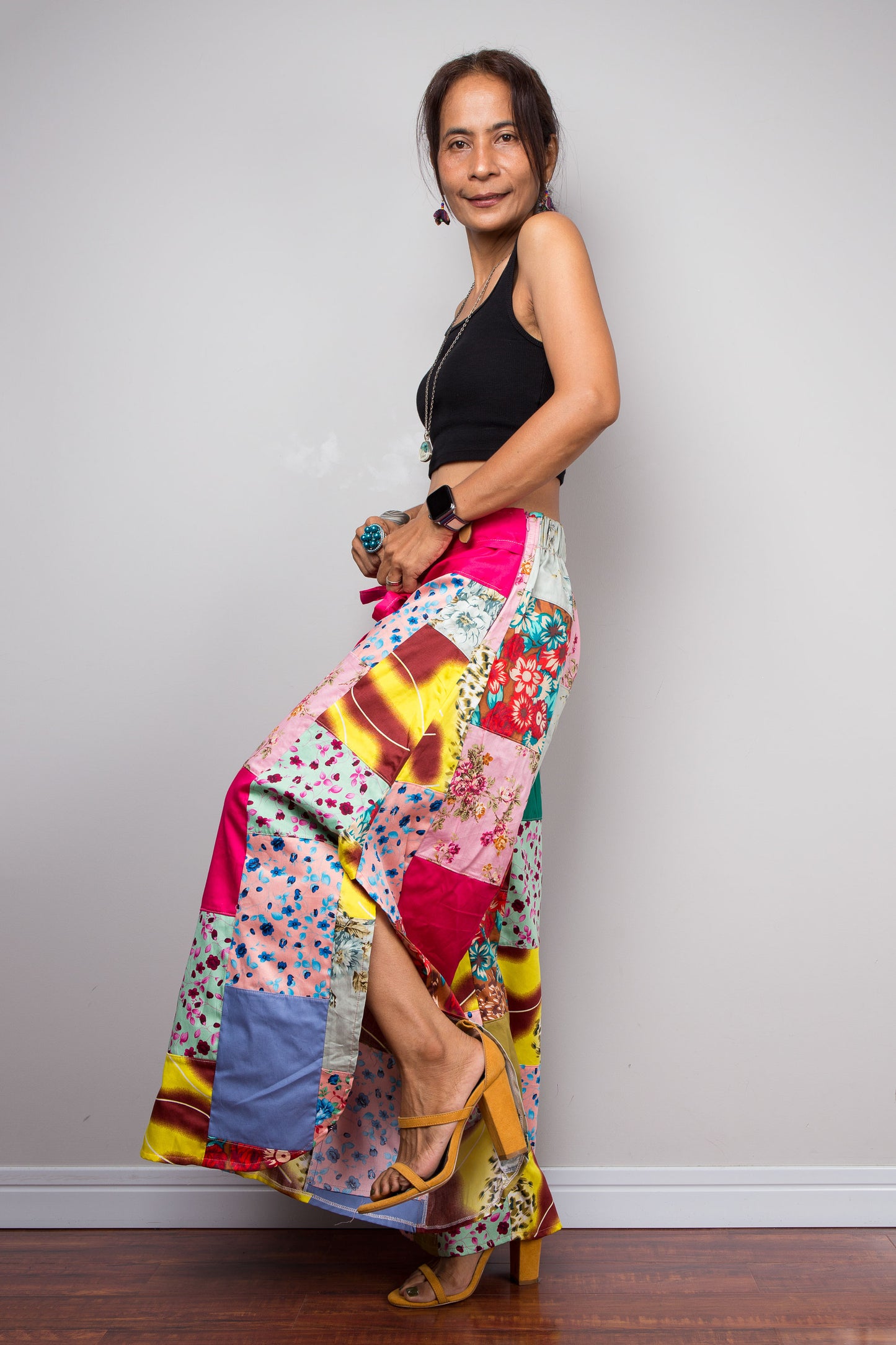 Side-facing view of a model wearing the Nuichan pastel patchwork wide leg palazzo pants, showcasing the pink self-tie sash waistband, full ultra-wide leg silhouette, and hand-sewn quilt-style patchwork construction in mixed cotton satin pastel prints including mint, blush pink, teal, yellow satin and floral panels, styled with a black crop top