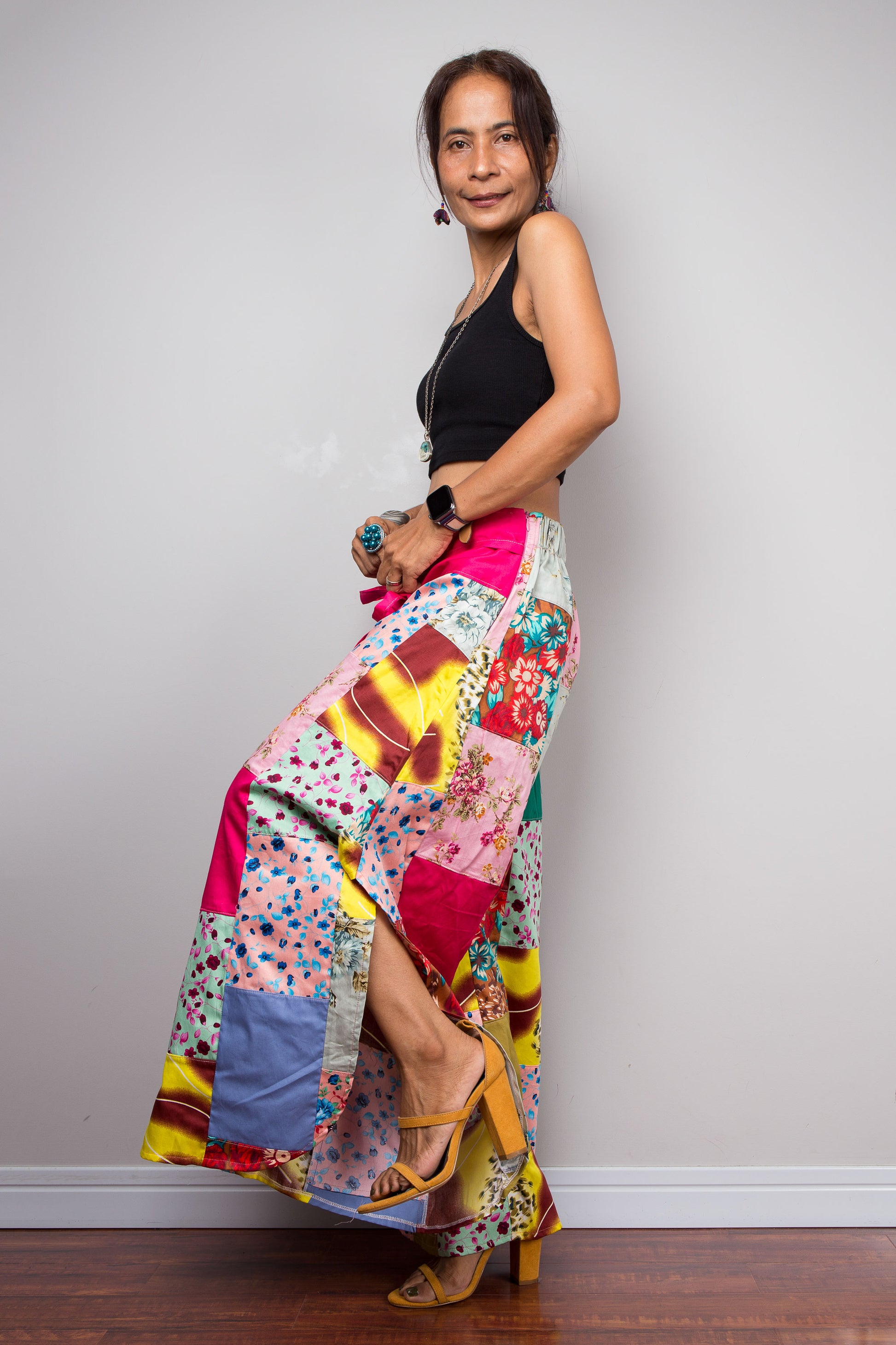Side-facing view of a model wearing the Nuichan pastel patchwork wide leg palazzo pants, showcasing the pink self-tie sash waistband, full ultra-wide leg silhouette, and hand-sewn quilt-style patchwork construction in mixed cotton satin pastel prints including mint, blush pink, teal, yellow satin and floral panels, styled with a black crop top