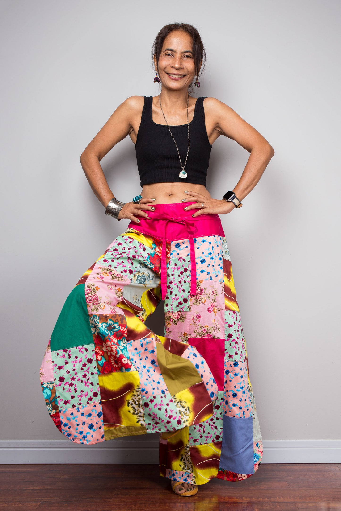 Front-facing view of a petite model wearing the Nuichan pastel patchwork wide leg palazzo pants, showcasing the vivid fuchsia pink self-tie sash waistband, full ultra-wide leg silhouette, and hand-sewn quilt-style patchwork construction in mixed cotton satin pastel prints including mint, blush pink, teal, yellow satin and floral panels, styled with a black crop top