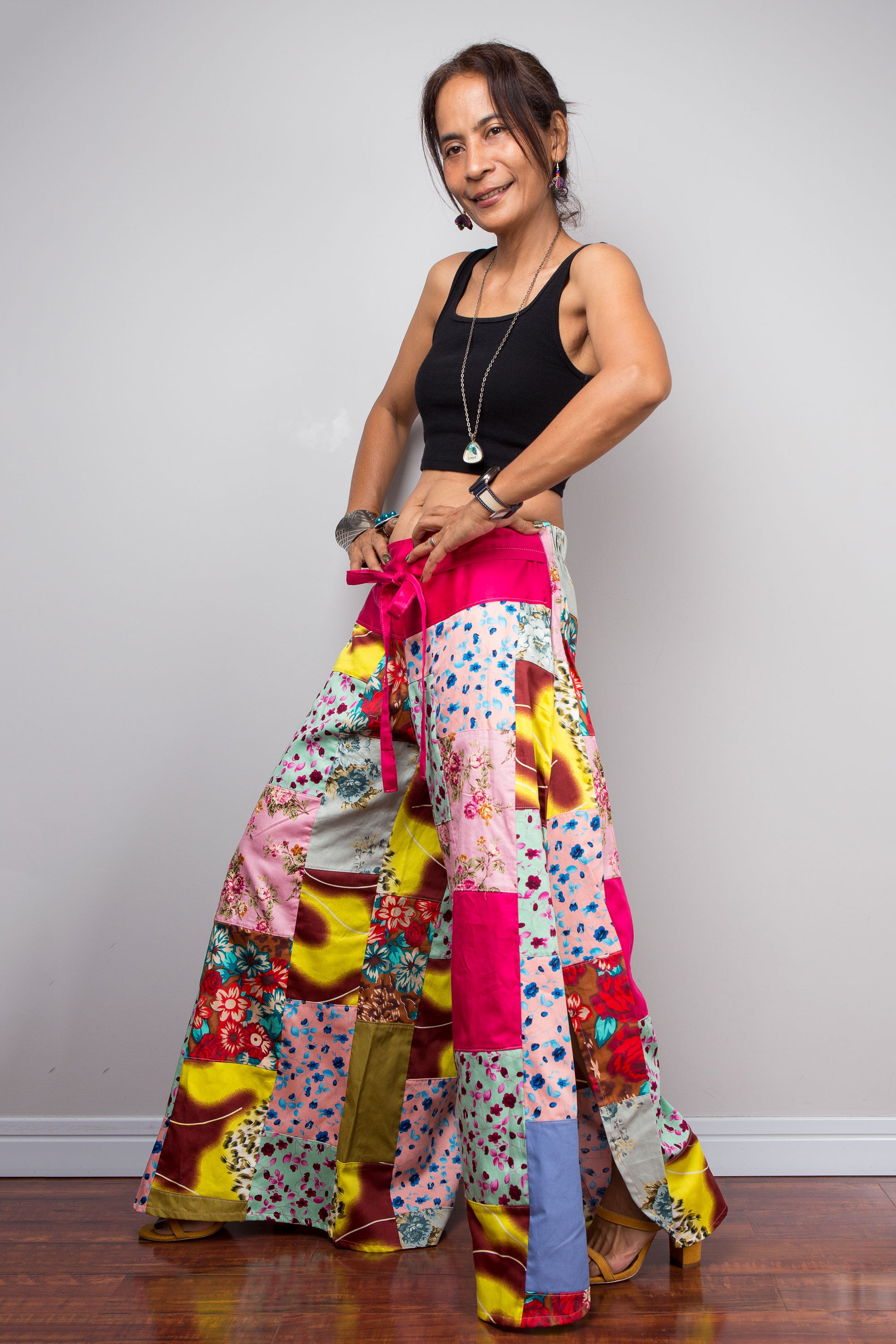 Side-facing view of a petite female model wearing the Nuichan pastel patchwork wide leg palazzo pants, showcasing the hot pink self-tie sash waistband, full ultra-wide leg silhouette, and hand-sewn quilt-style patchwork construction in mixed cotton satin pastel prints including mint, blush pink, teal, yellow satin and floral panels, styled with a black crop top