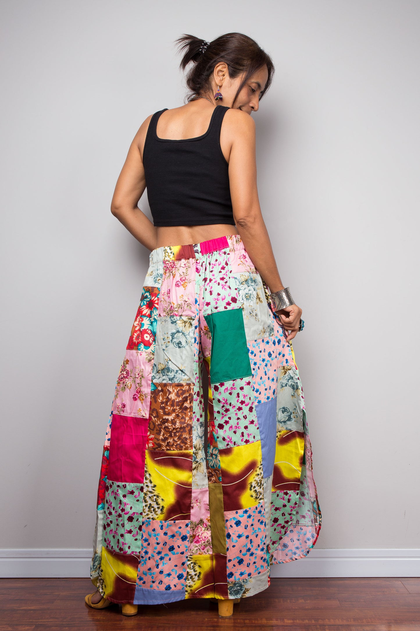 Rear view of a model wearing the Nuichan pastel patchwork wide leg palazzo pants, full ultra-wide leg silhouette, and hand-sewn quilt-style patchwork construction in mixed cotton satin pastel prints including mint, blush pink, teal, yellow satin and floral panels, styled with a black crop top