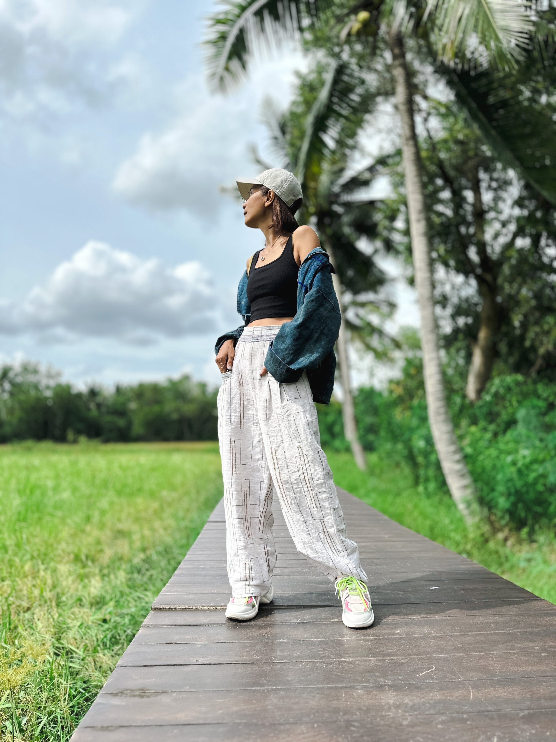 Model wearing handwoven hemp harem pants with a natural beige base and multicolored vertical stripes in blue, red, and yellow. These boho harem pants, paired with a black crop top and denim jacket, are styled on a wooden boardwalk in a lush green setting.
