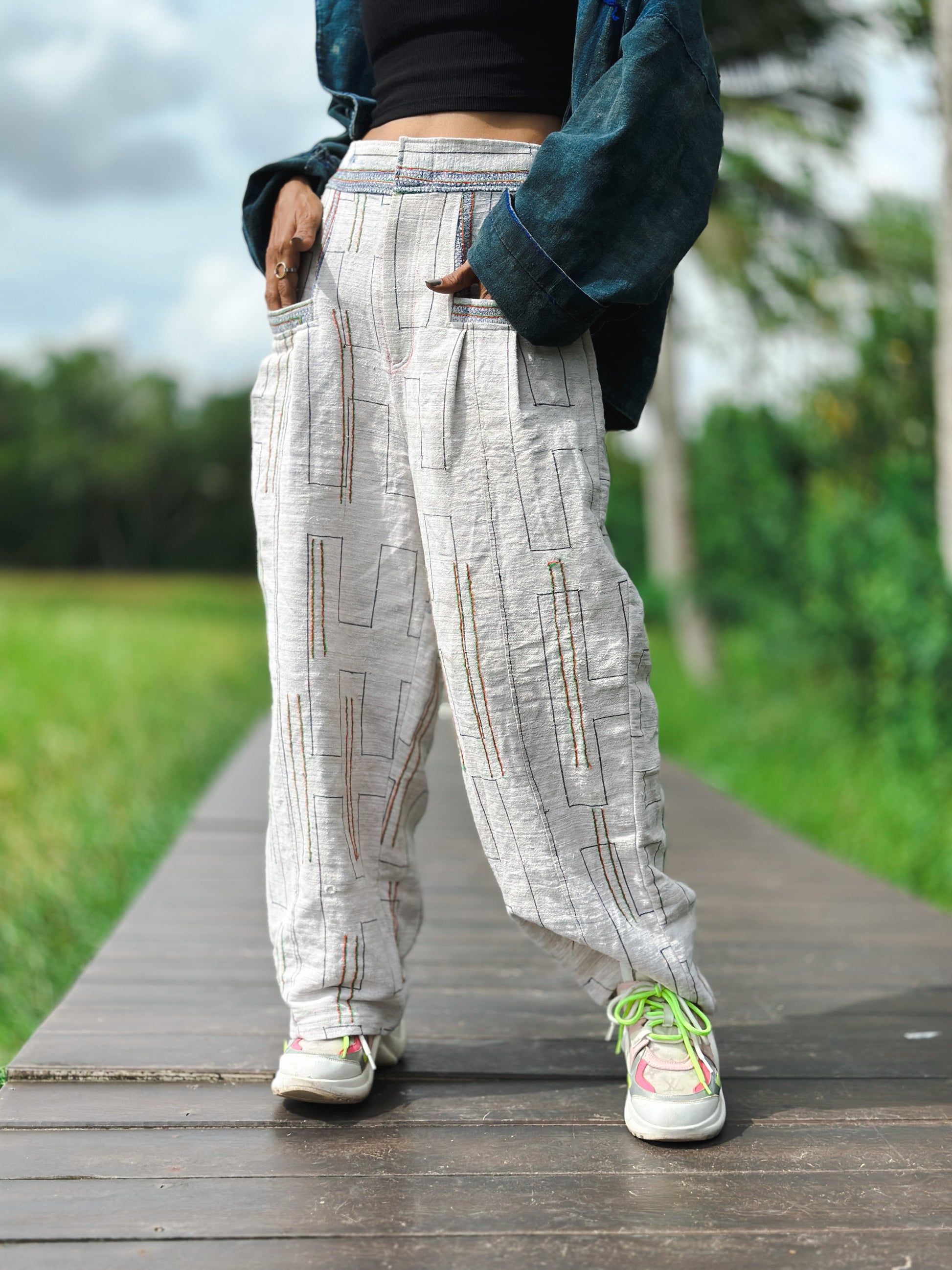 Model in handwoven hemp harem pants featuring a natural beige tone with multicolored stripes in blue, red, and yellow. These striped boho pants, styled with a denim jacket, create an earthy aesthetic on a wooden boardwalk with a green field in the background.