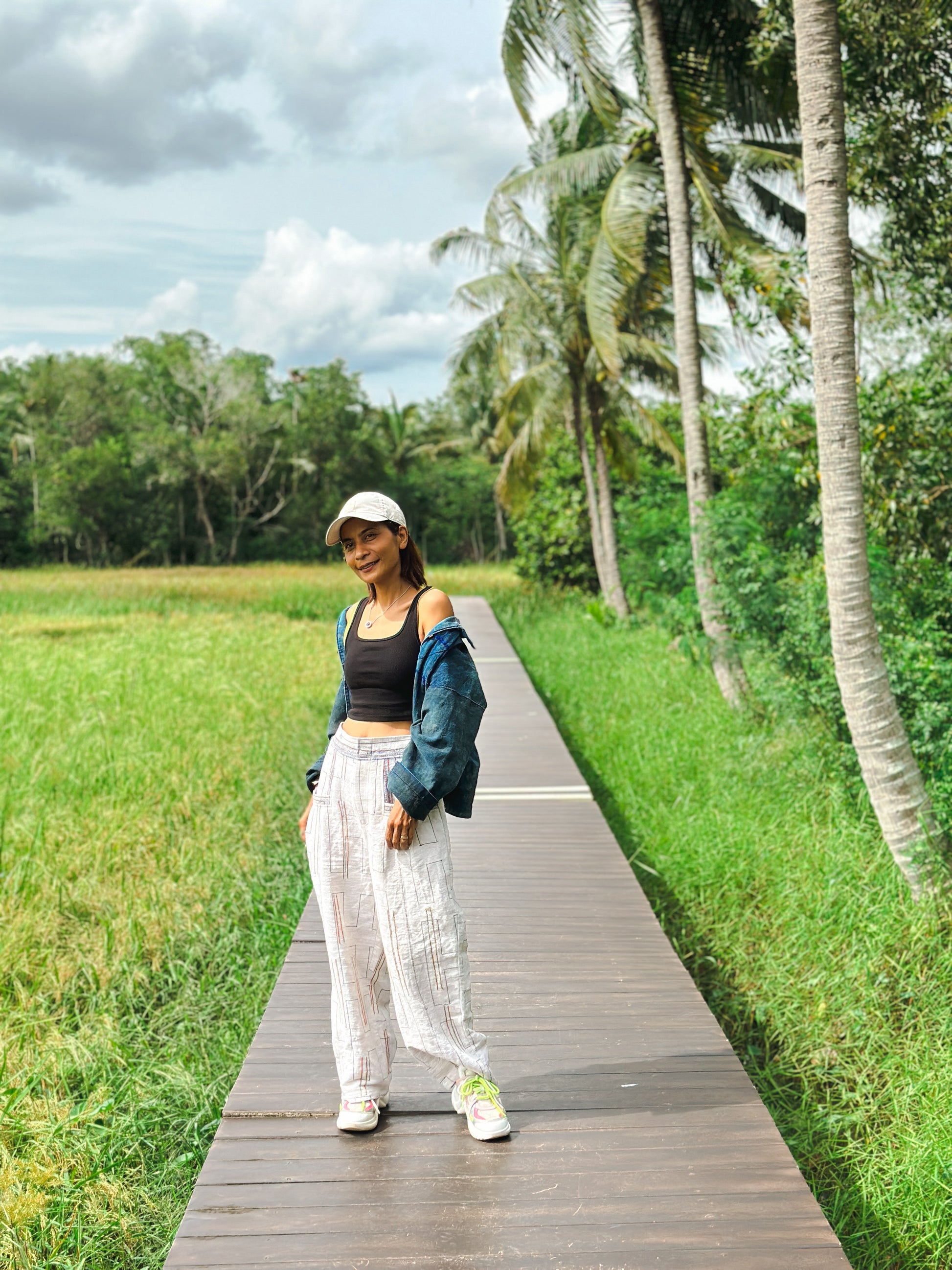 Handwoven hemp harem pants in natural beige with multicolored stripes, featuring a relaxed high-waisted fit and gathered cuffs. These comfortable boho pants, styled with a denim jacket, are showcased on a wooden boardwalk with a tropical green background.