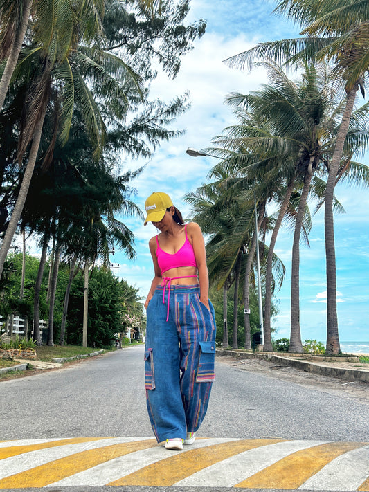 A person standing on a crosswalk wearing blue handwoven hemp cargo pants with a colorful top and a yellow hat, with palm trees and a clear blue sky in the background.