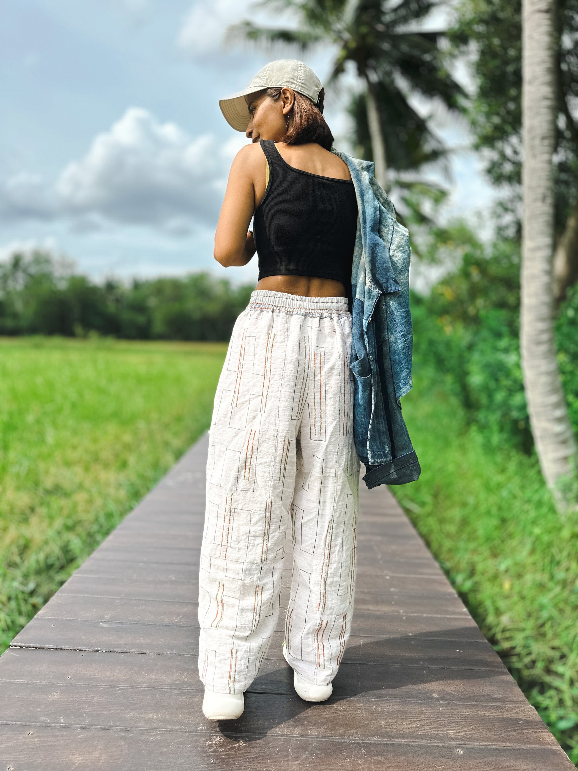 Woman styling handwoven hemp harem pants in natural beige with multicolored stripes, paired with a black crop top, denim jacket, and colorful sneakers. These sustainable boho pants, photographed on a wooden boardwalk, are accessorized with a cap for a chic look.
