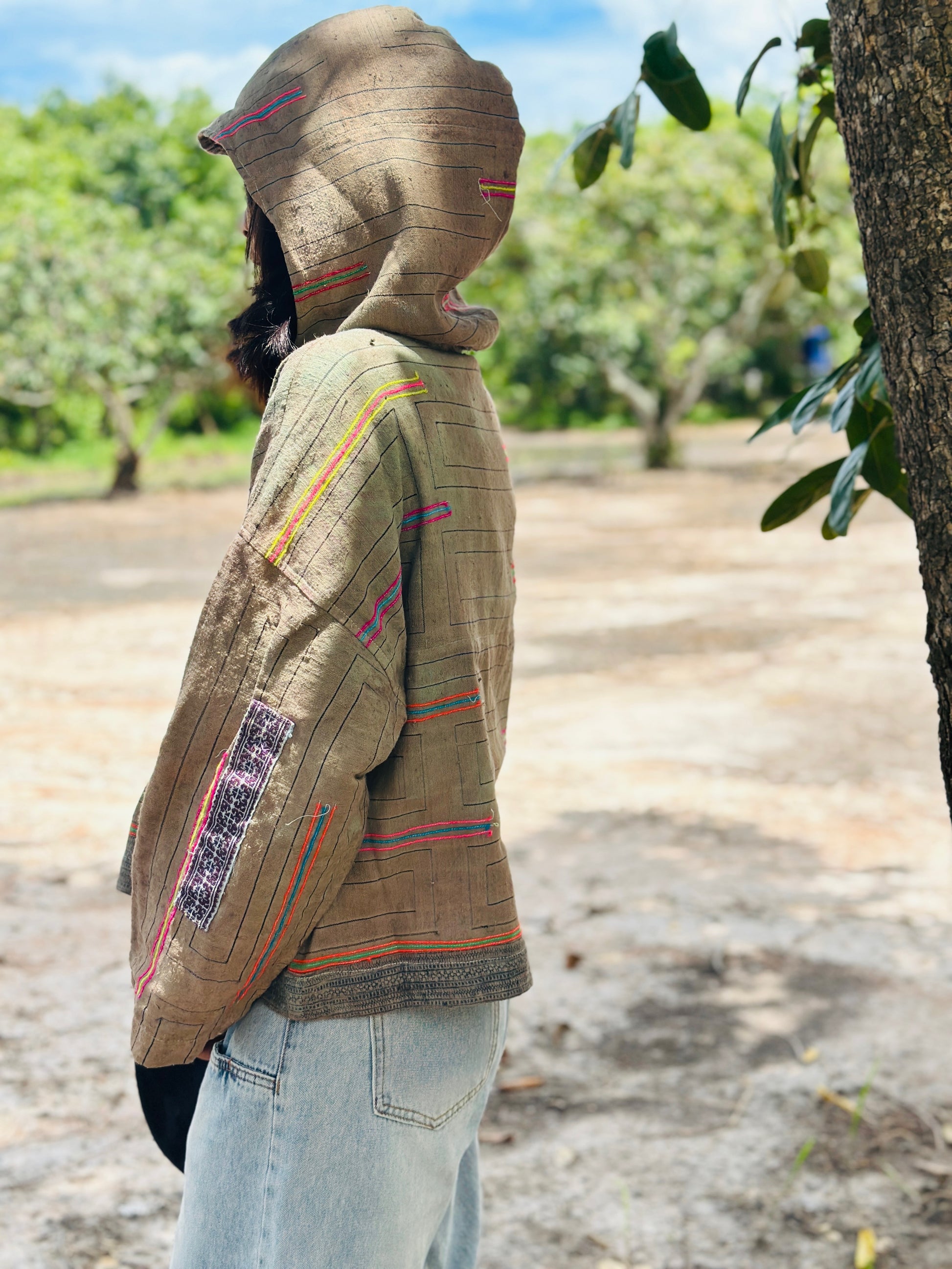 Person wearing a traditional woven jacket in an outdoor setting with trees and a clear sky.