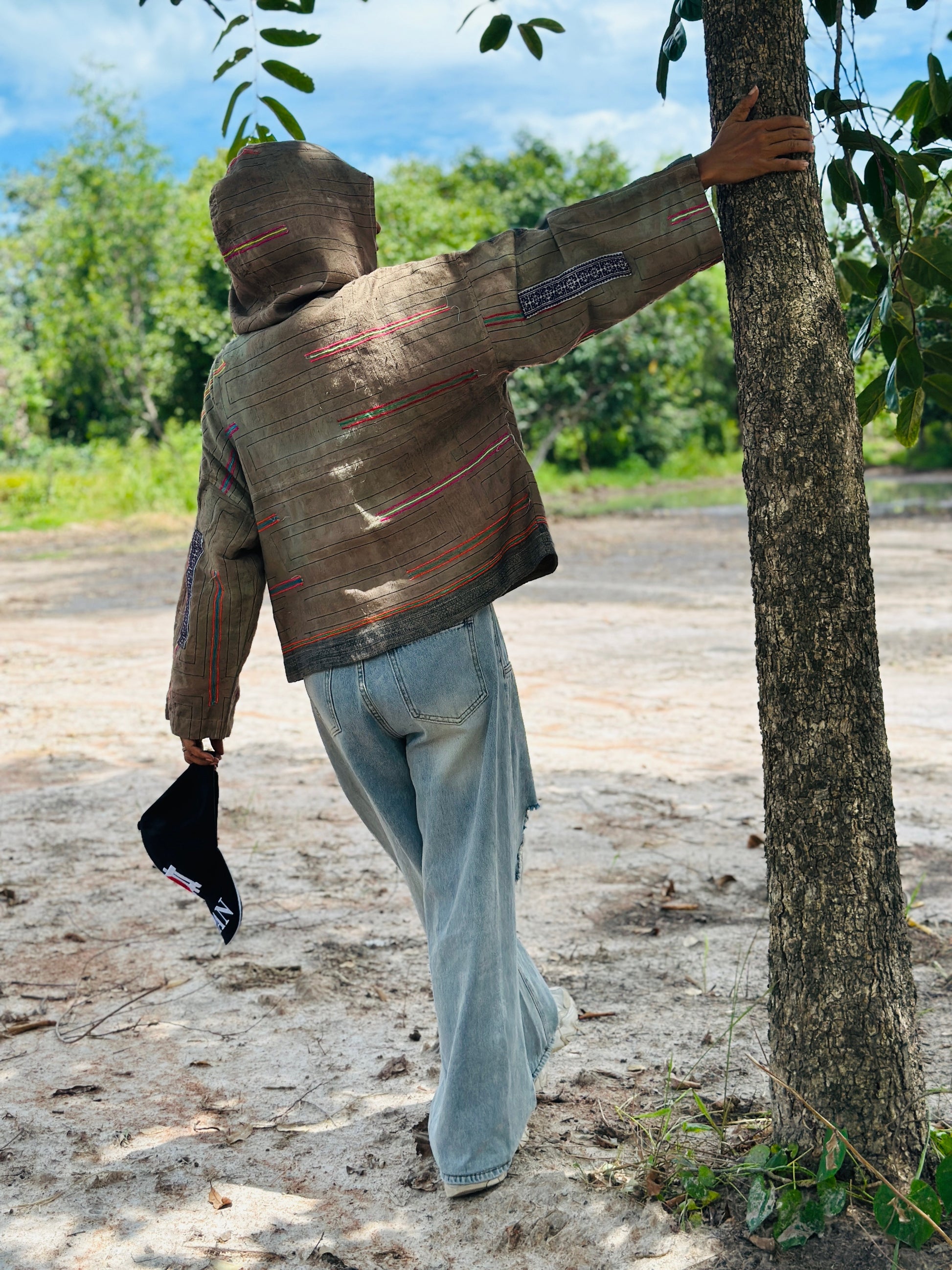 Person wearing a brown hooded jacket and light blue jeans leaning against a tree outdoors.