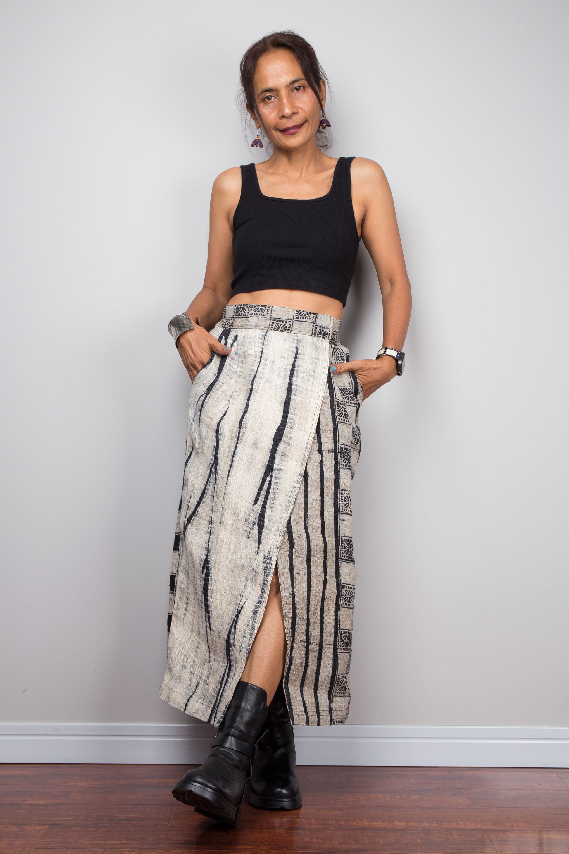 Front view of a petite female model wearing the Nuichan handwoven hemp wrap skirt with hands in side pockets, displaying the wrap front construction with flat metal hook and bar closures, Japanese shibori vertical lightning stripe pattern on the front panel, hand-drawn batik checkerboard side panel, front split opening, and hand-drawn batik geometric waistband