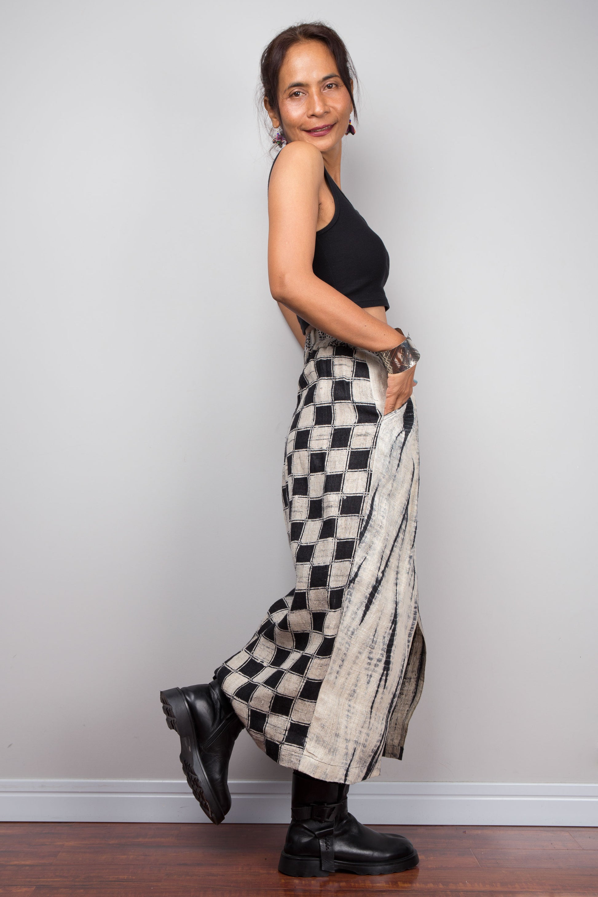 A woman posing in a black sleeveless crop top and a long, wide-leg skirt featuring a black and beige checkerboard pattern with a textured beige stripe on the side, paired with black boots, against a plain light gray wall.