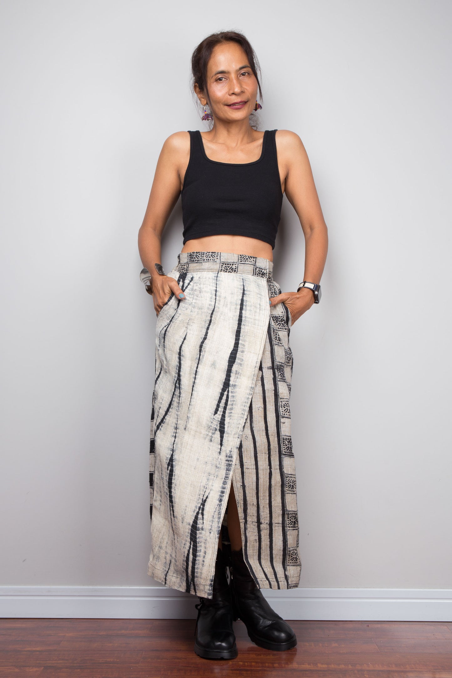 Front-facing view of a petite model wearing the Nuichan handwoven hemp wrap skirt with hands in side pockets, displaying the wrap front construction with flat metal hook and bar closures, Japanese shibori vertical lightning stripe pattern on the front panel, hand-drawn batik checkerboard side panel, front split opening, and hand-drawn batik geometric waistband