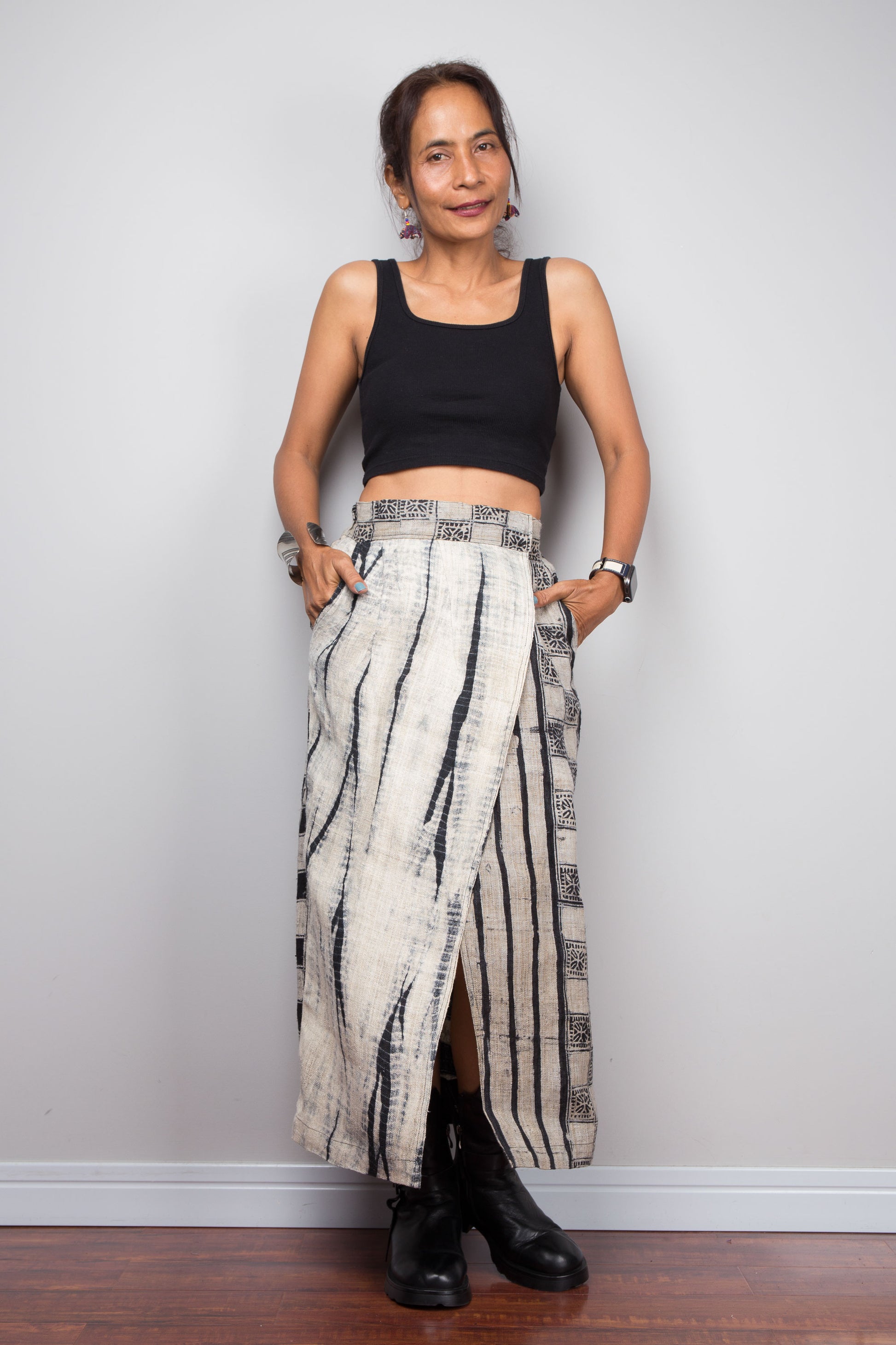 Front-facing view of a petite model wearing the Nuichan handwoven hemp wrap skirt with hands in side pockets, displaying the wrap front construction with flat metal hook and bar closures, Japanese shibori vertical lightning stripe pattern on the front panel, hand-drawn batik checkerboard side panel, front split opening, and hand-drawn batik geometric waistband