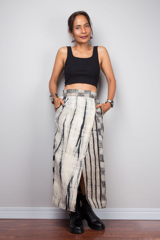 Front-facing view of a petite model wearing the Nuichan handwoven hemp wrap skirt with hands in side pockets, displaying the wrap front construction with flat metal hook and bar closures, Japanese shibori vertical lightning stripe pattern on the front panel, hand-drawn batik checkerboard side panel, front split opening, and hand-drawn batik geometric waistband