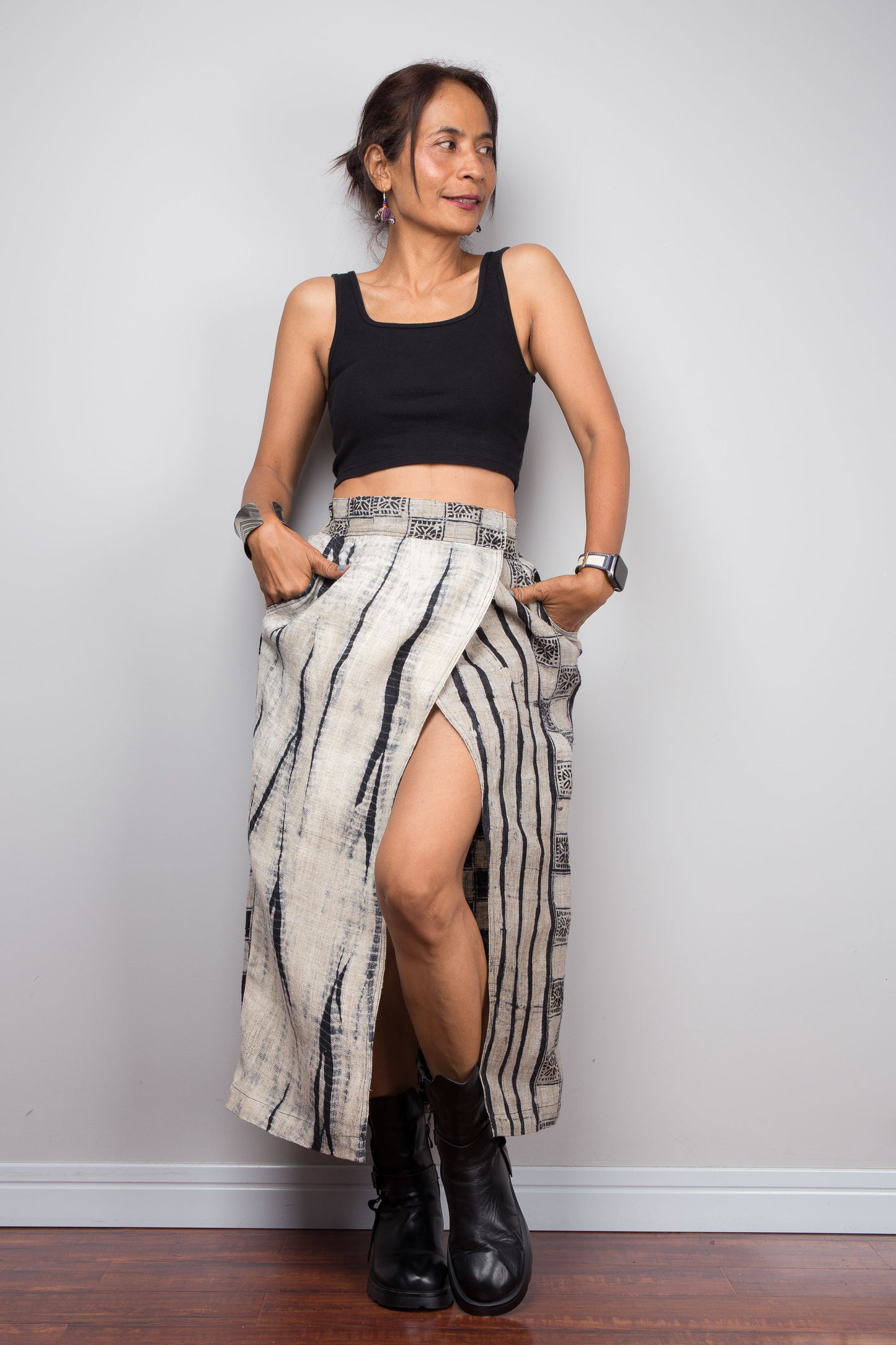 Front-facing view of a model wearing the Nuichan handwoven hemp petite wrap skirt with hands in side pockets, displaying the wrap front construction with flat metal hook and bar closures, Japanese shibori vertical lightning stripe pattern on the front panel, hand-drawn batik checkerboard side panel, front split opening, and hand-drawn batik geometric waistband