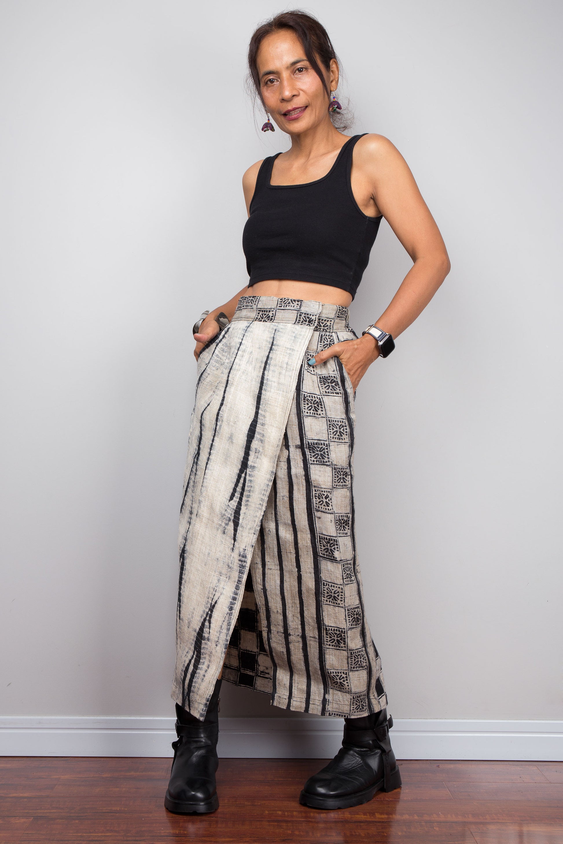 Front view of a model wearing the Nuichan handwoven hemp wrap skirt with hands in side pockets, displaying the Japanese shibori vertical lightning stripe pattern on the front panel, hand-drawn batik checkerboard side panel, front split opening, and hand-drawn batik geometric waistband