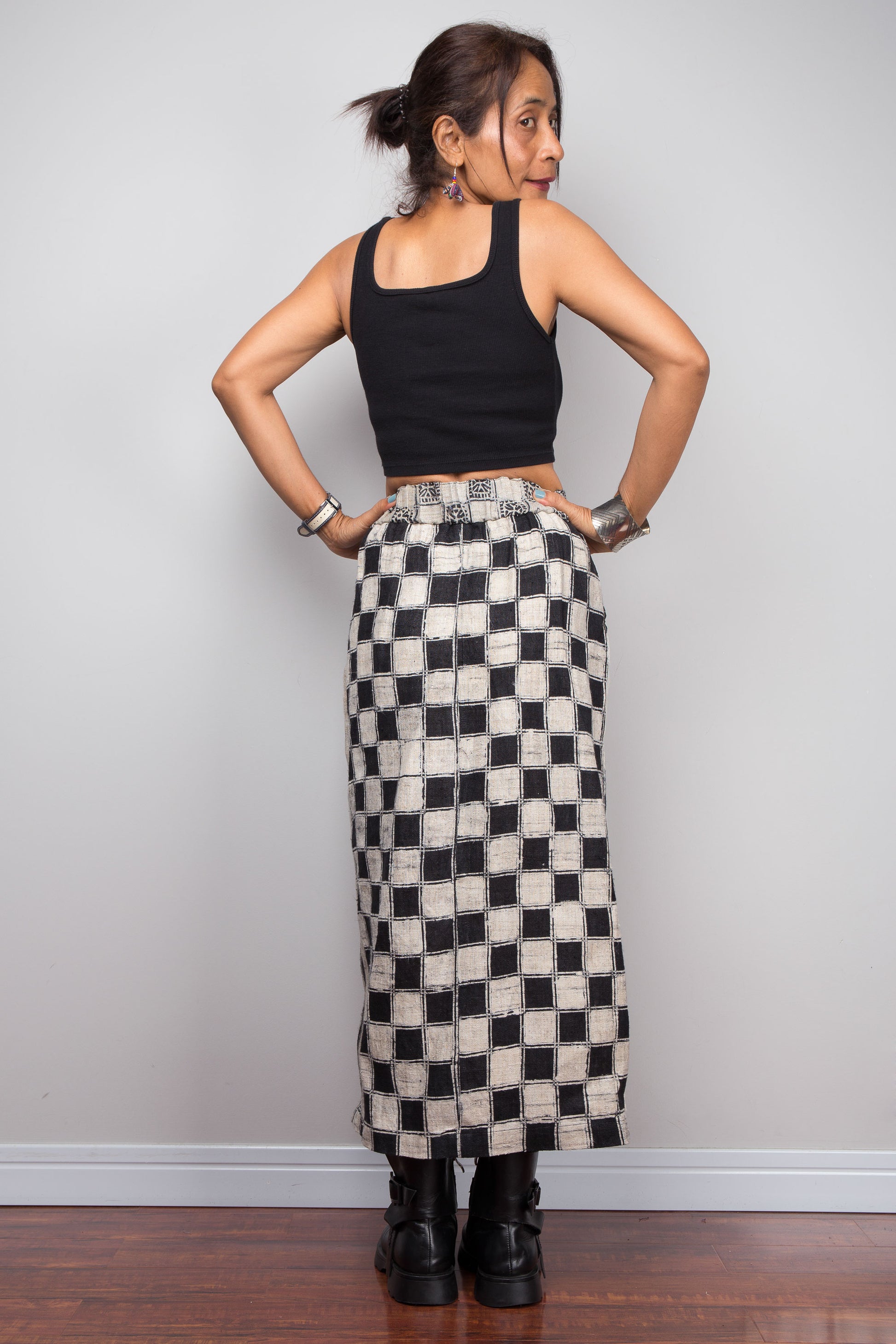 Rear view of a model wearing the Nuichan handwoven hemp wrap split skirt, showcasing the bold hand-drawn candle batik resist checkerboard pattern on the side panel in deep black and natural ecru hemp, elastic waistband with hand-drawn batik geometric repeat, and mid-calf length wrap silhouette styled with a black crop top and chunky ankle boots