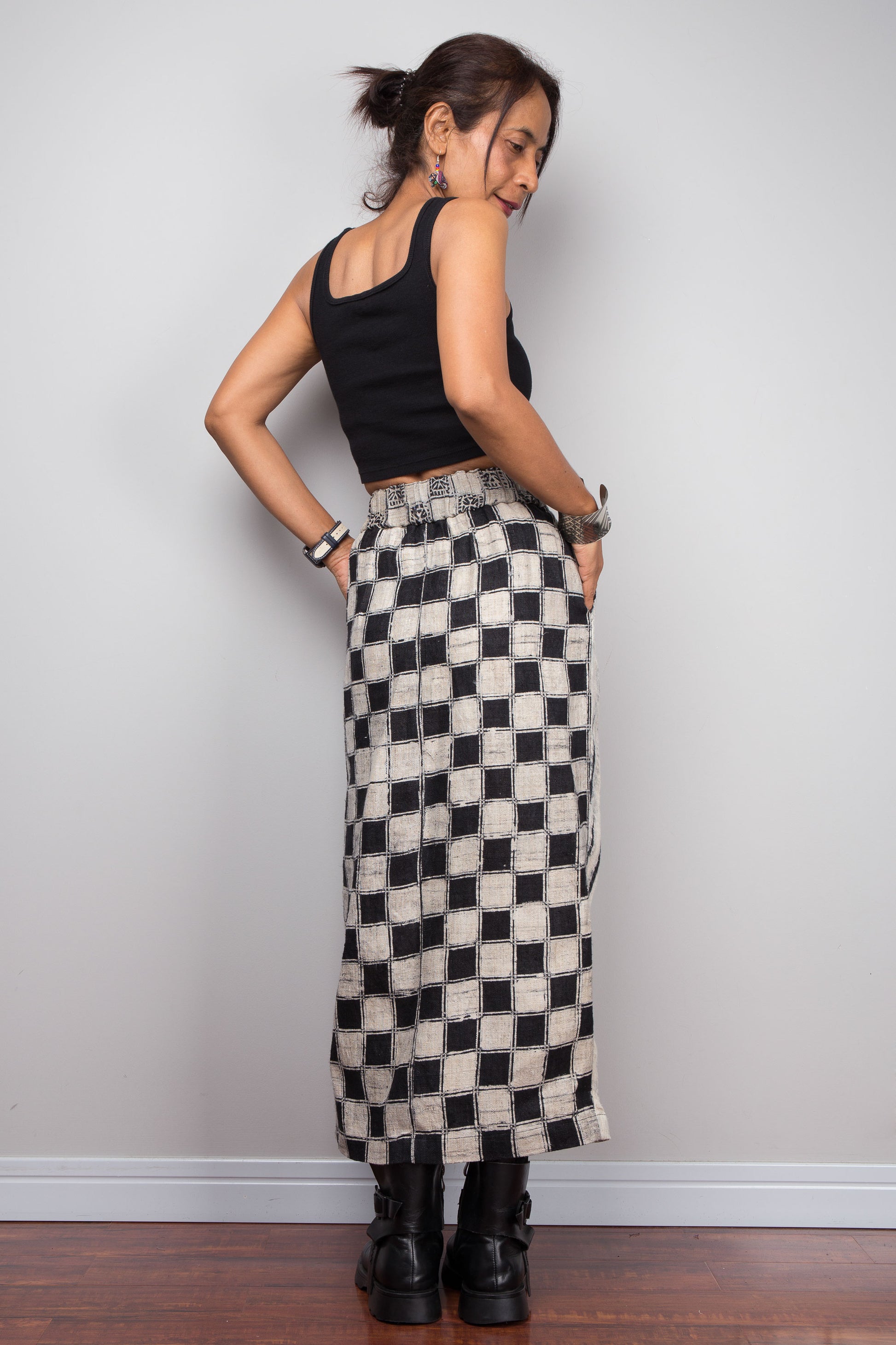 Back view of a model wearing the Nuichan handwoven hemp wrap petite skirt with hands in side pockets, displaying the wrap front construction with flat metal hook and bar closures, Japanese shibori vertical lightning stripe pattern on the front panel, hand-drawn batik checkerboard side panel, front split opening, and hand-drawn batik geometric waistband