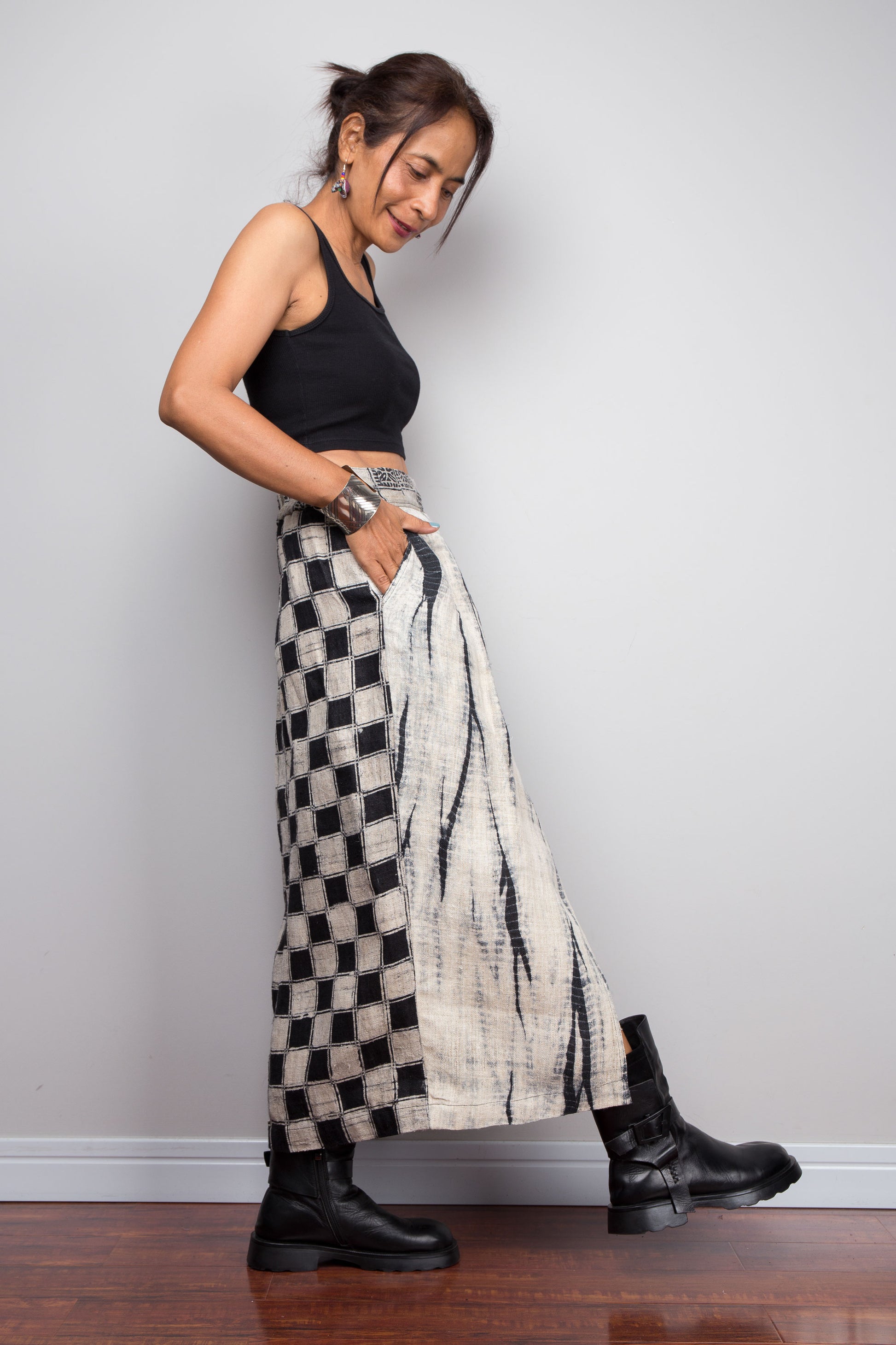 Side view of a female model wearing the Nuichan handwoven hemp wrap petite skirt with hands in side pockets, displaying the wrap front construction with flat metal hook and bar closures, Japanese shibori vertical lightning stripe pattern on the front panel, hand-drawn batik checkerboard side panel, front split opening, and hand-drawn batik geometric waistband