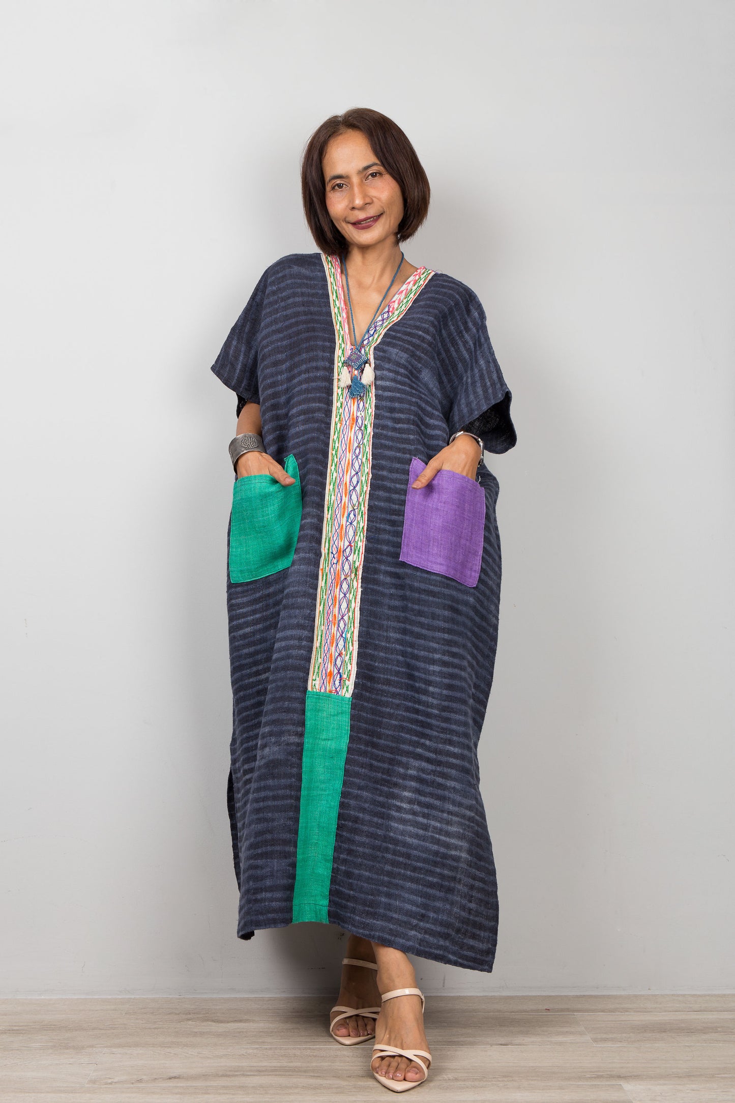 Woman wearing a long, dark blue kaftan dress with colorful pockets against a plain background