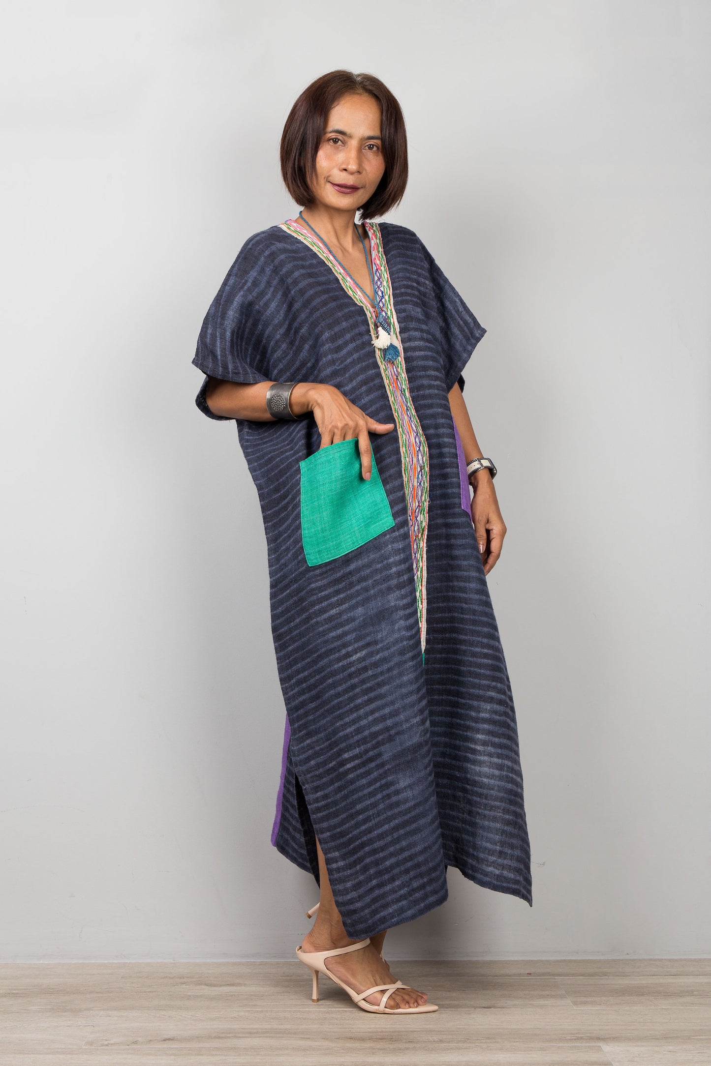 Woman wearing a long, dark blue striped hemp kaftan dress with a green pocket against a plain background