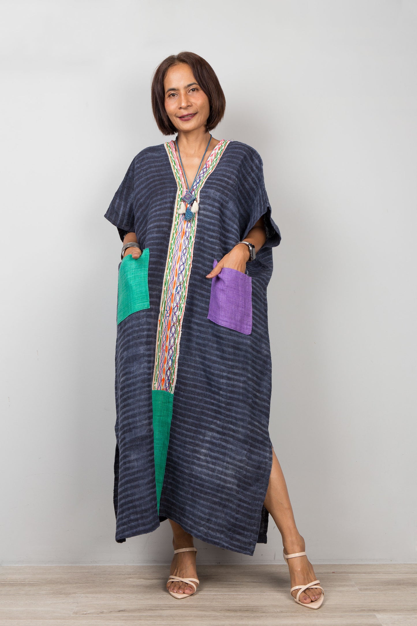 Woman wearing a long, dark blue hemp caftan dress with colorful pockets on a plain background