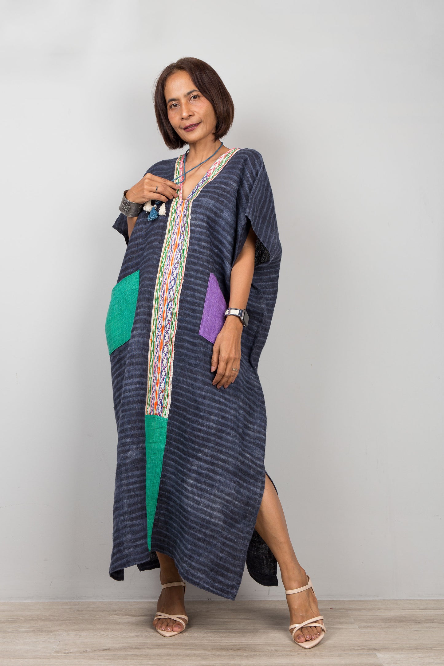 Woman wearing a long, dark blue hemp caftan with colorful pockets on a plain background
