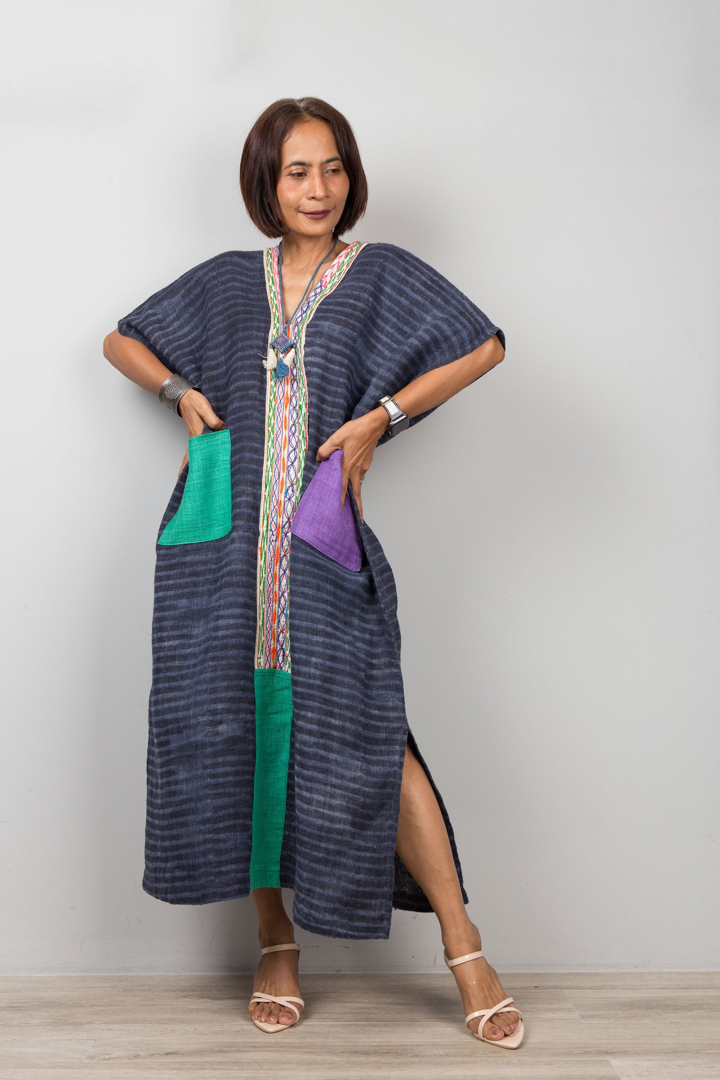 Woman wearing a long, dark blue kaftan dress with colorful pockets on a plain background.  Handwoven hemp caftan by Nuichan