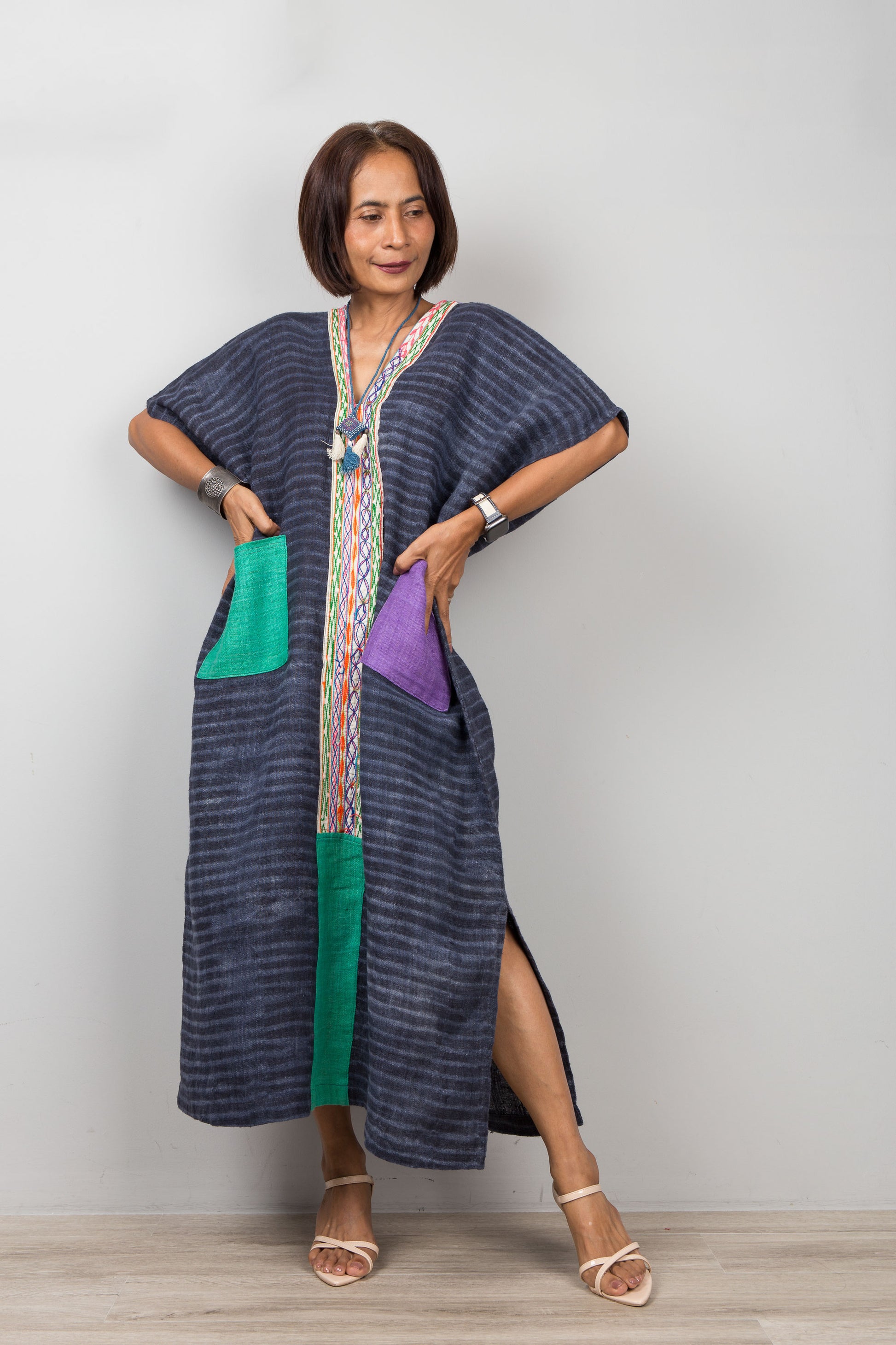 Woman wearing a long, dark blue kaftan dress with colorful pockets on a plain background.  Handwoven hemp caftan by Nuichan