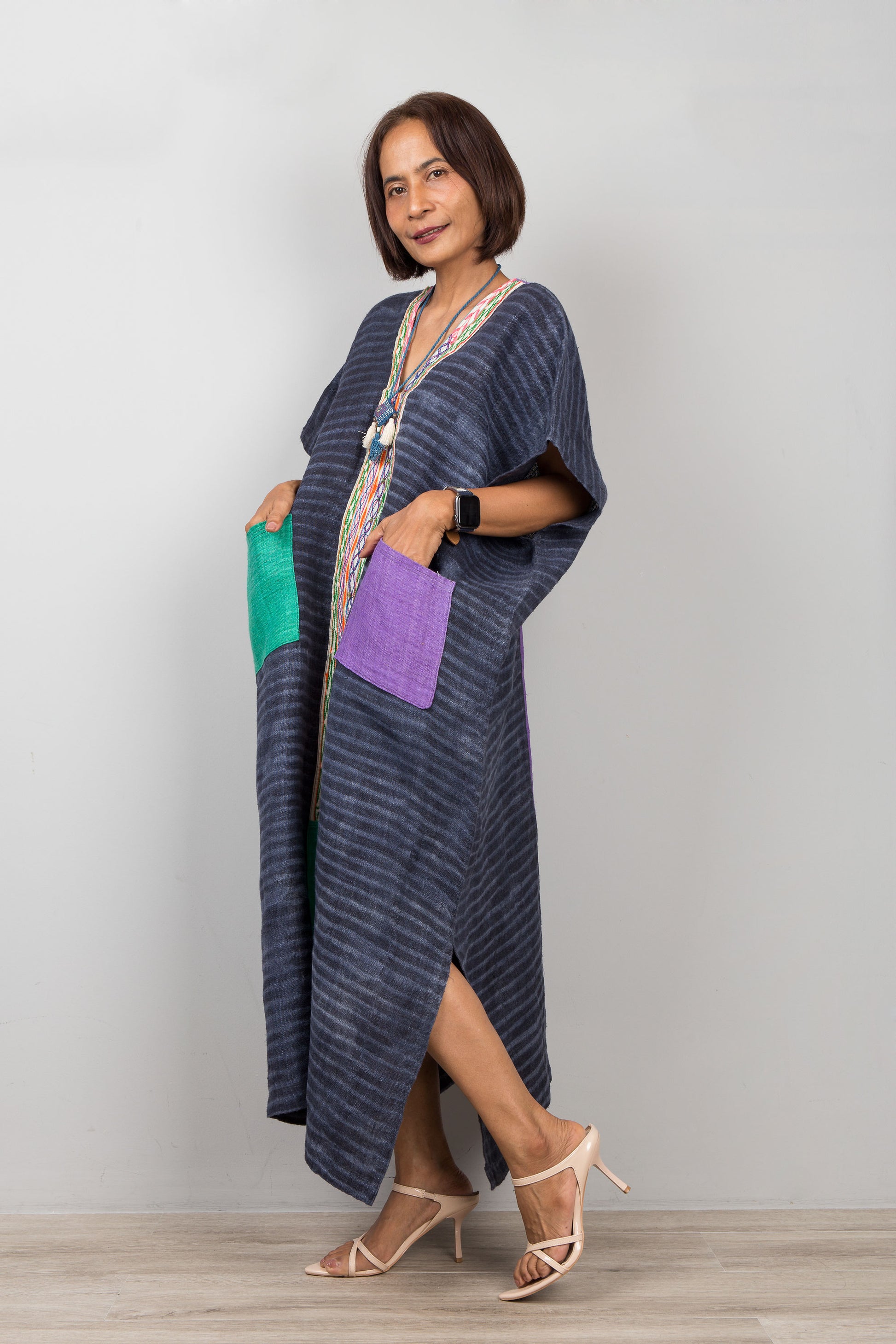 Woman wearing a long, dark blue hemp kaftan dress with colorful pockets against a plain background