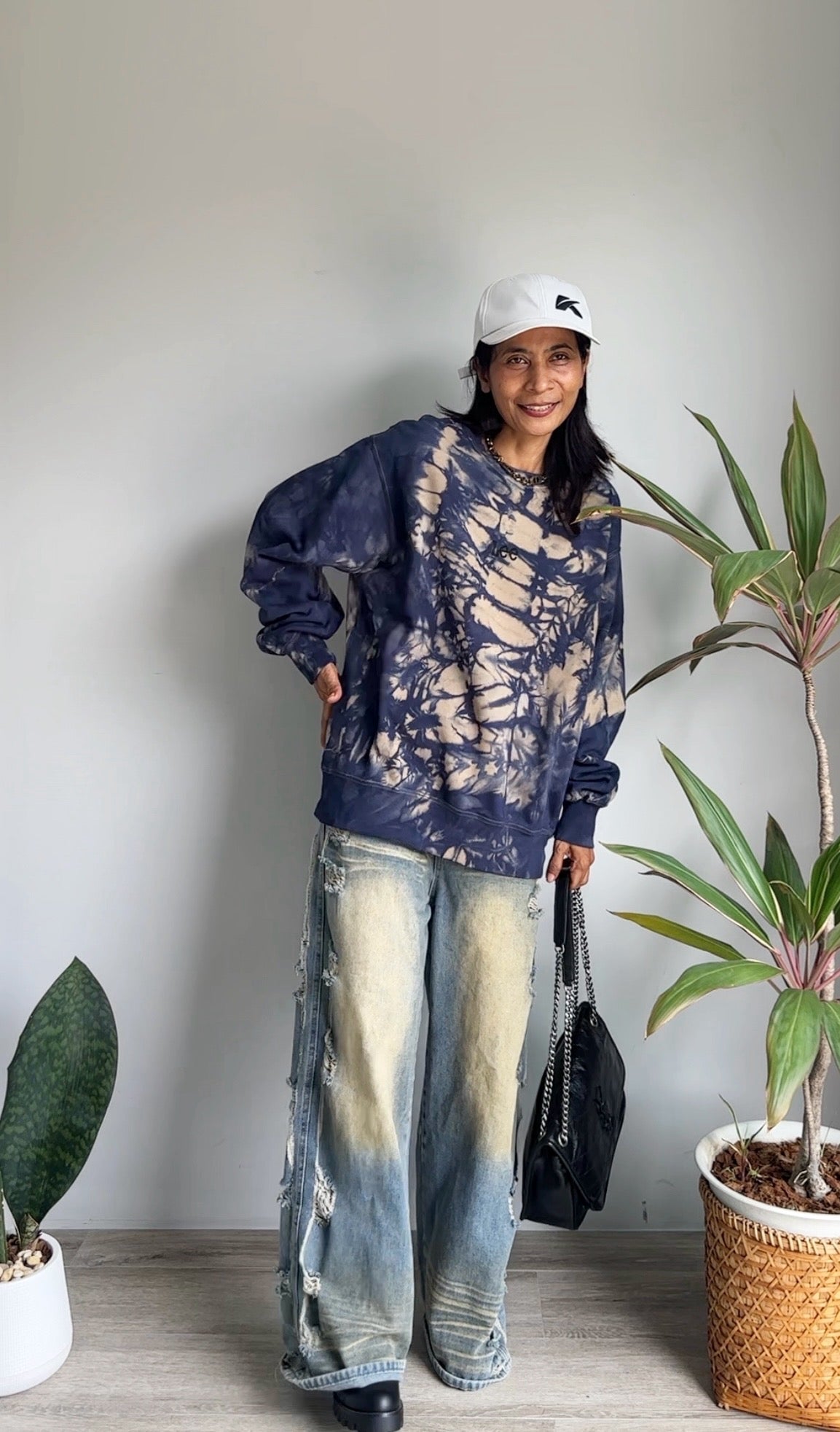 Person wearing a tie-dye sweatshirt and jeans standing indoors with plants around.