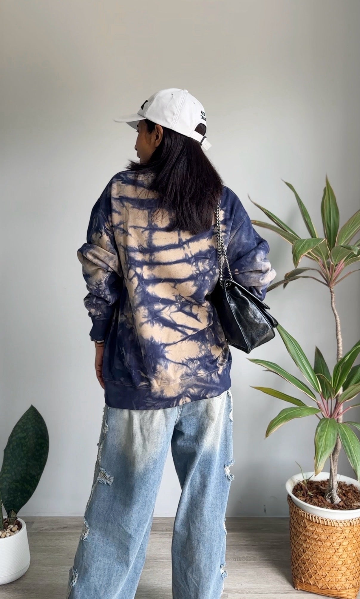 Person wearing a tie-dye shirt and jeans standing in a room with plants.